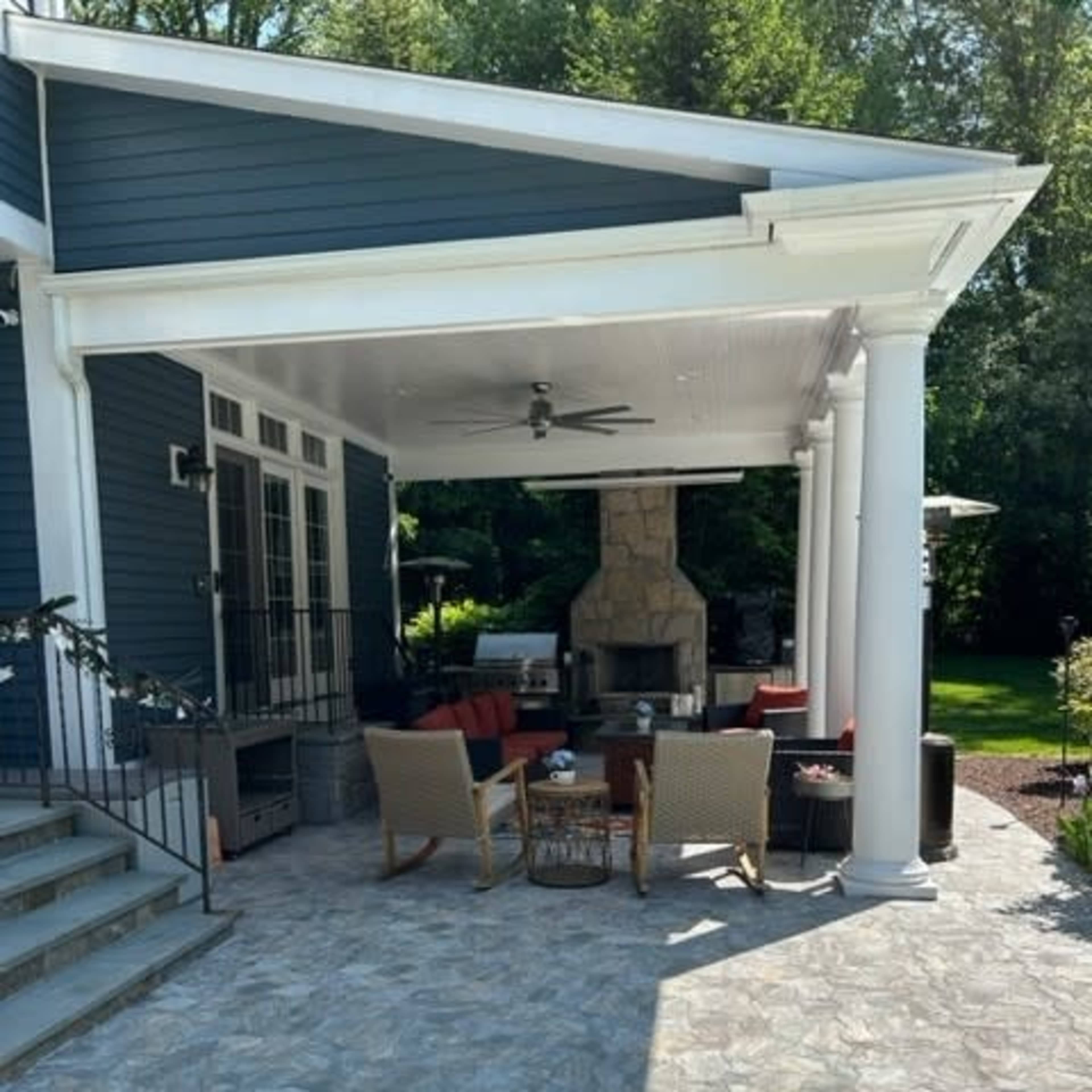 A patio area with woven chairs around a coffee table, featuring a stone fireplace and pillars, set against a green yard.