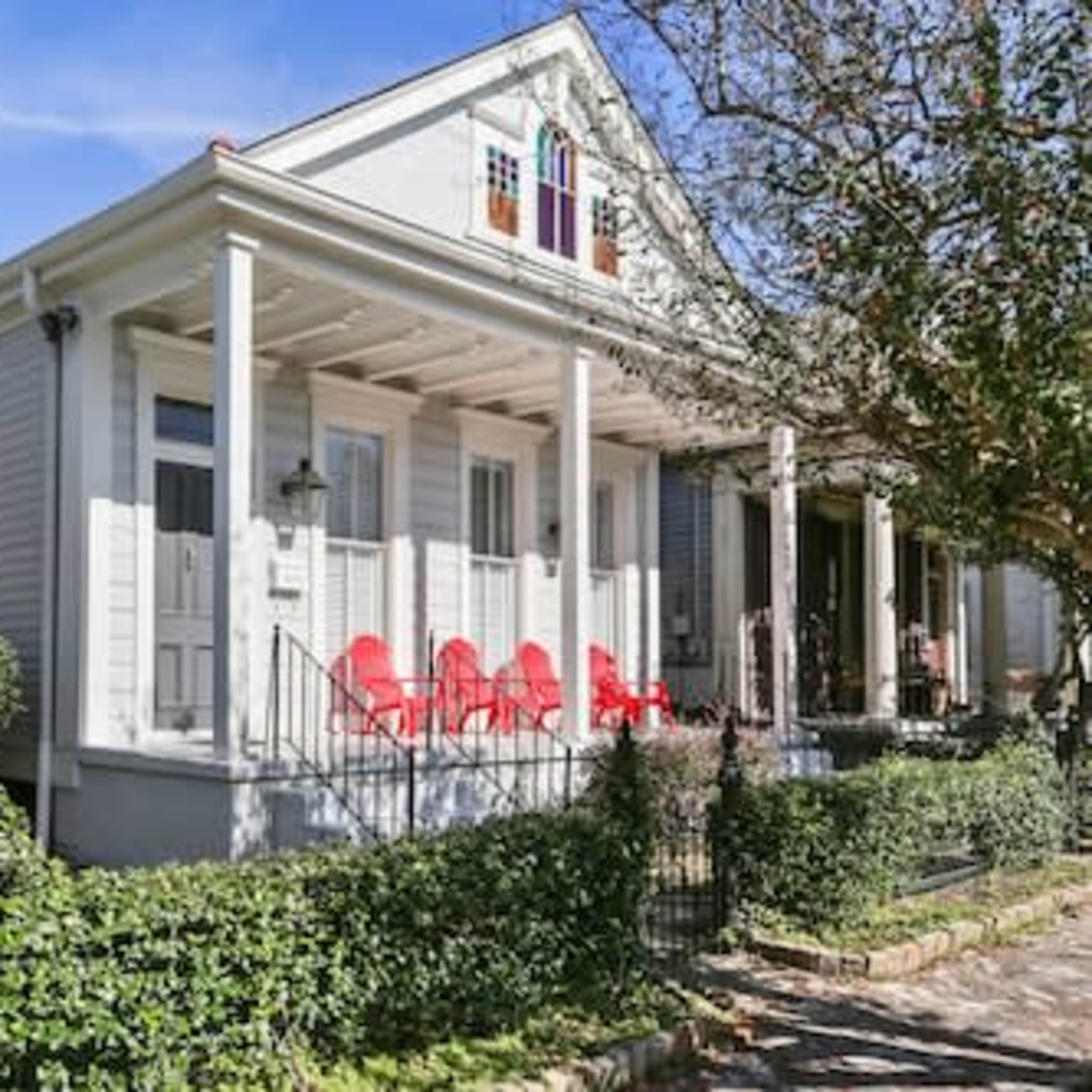 A light-colored house with a covered porch and red rocking chairs sits at the edge of a well-maintained garden.