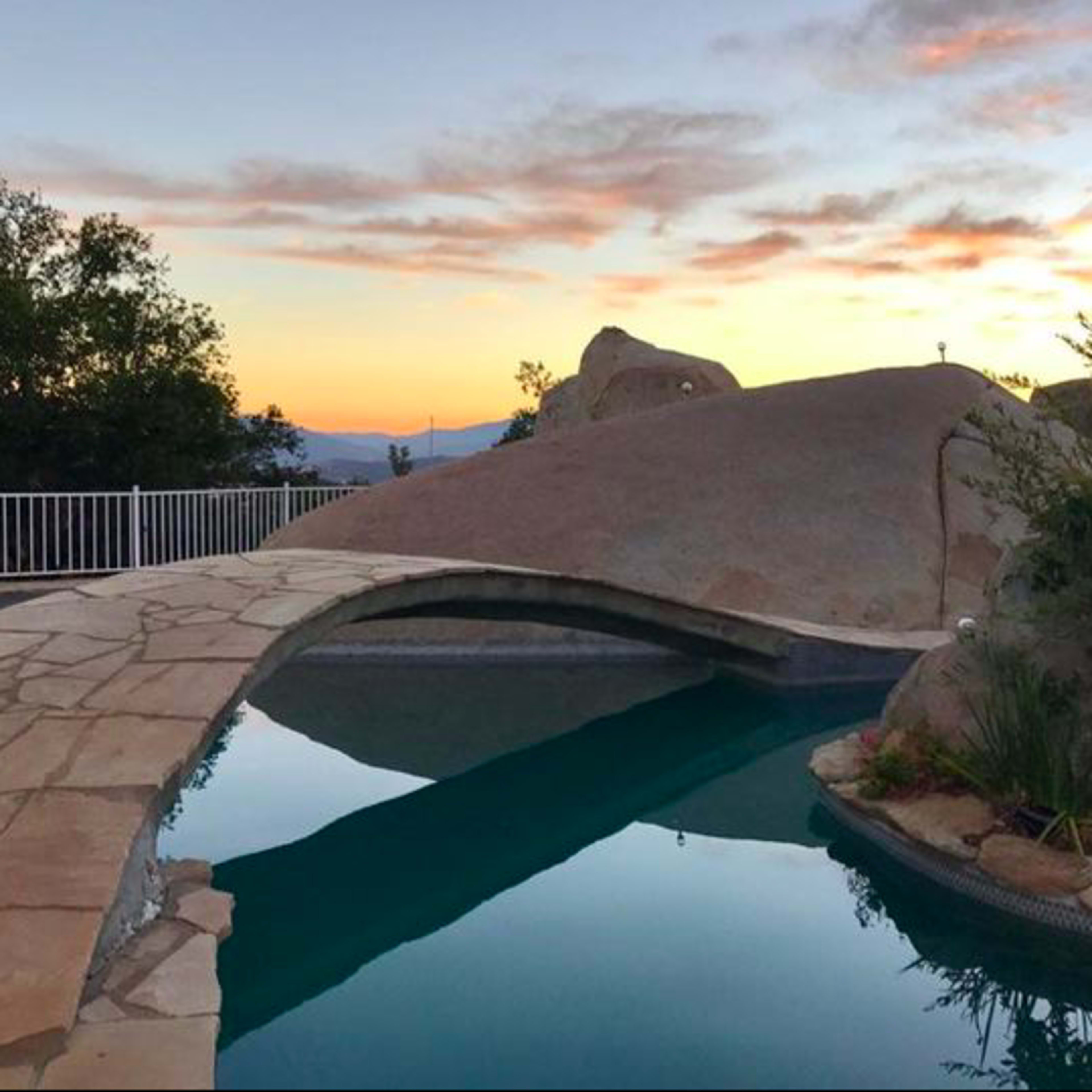 A stone pathway arches over a swimming pool, leading to a rocky landscape under a colorful sunset sky.