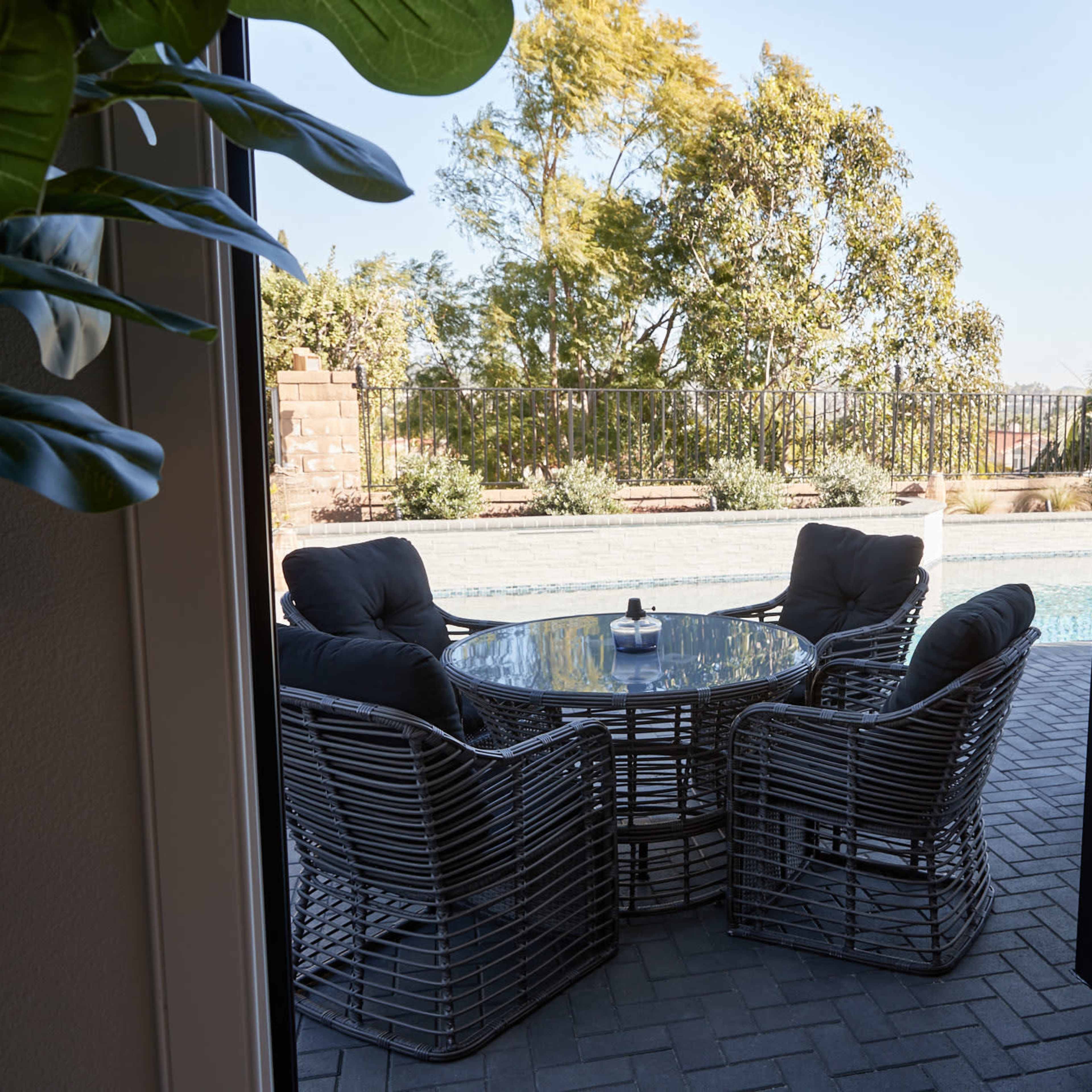 A circular glass table surrounded by four wicker chairs is positioned near a swimming pool, framed by a doorway with a view of greenery in the background.
