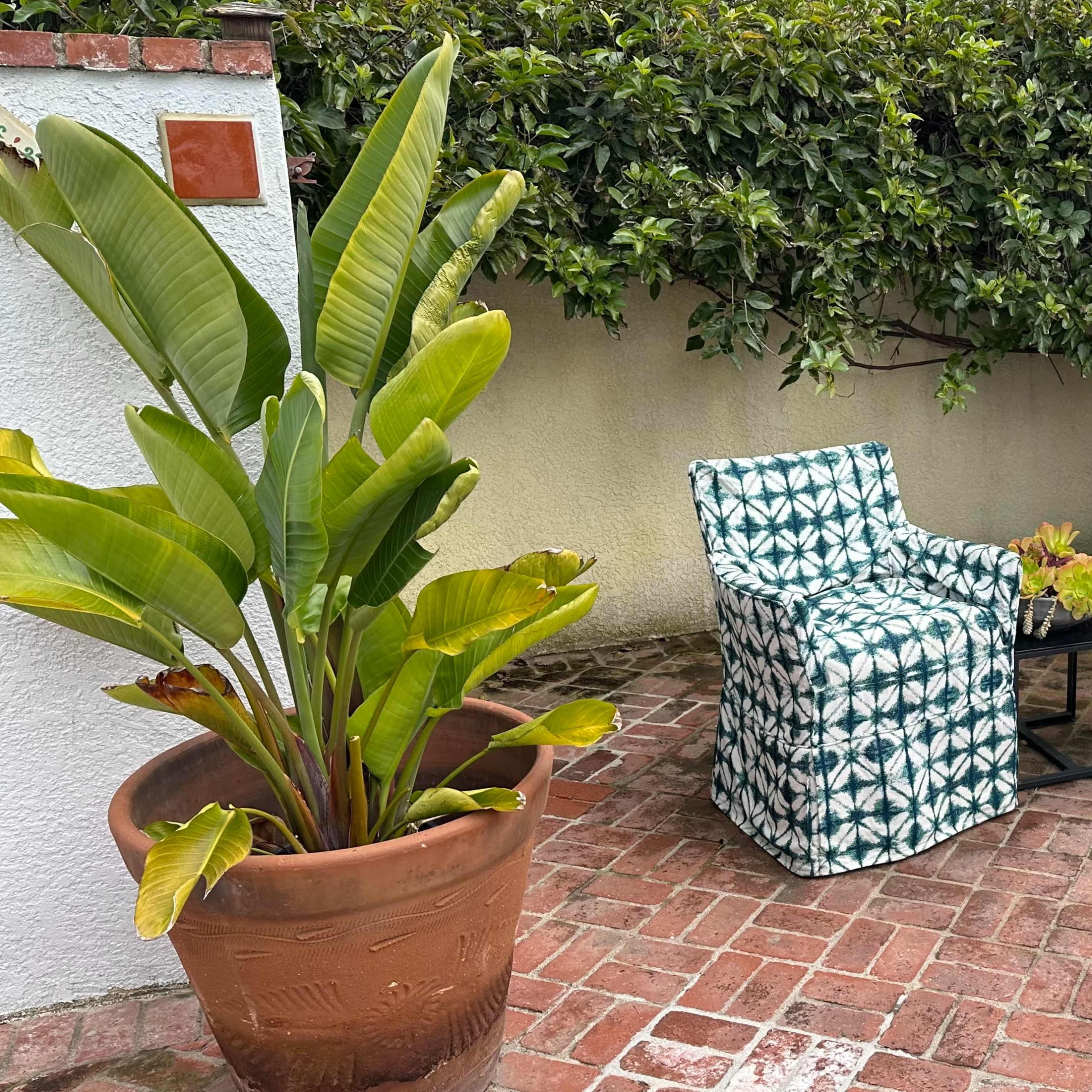 Two patterned chairs are placed next to a small table in a patio surrounded by large potted plants and a brick floor.