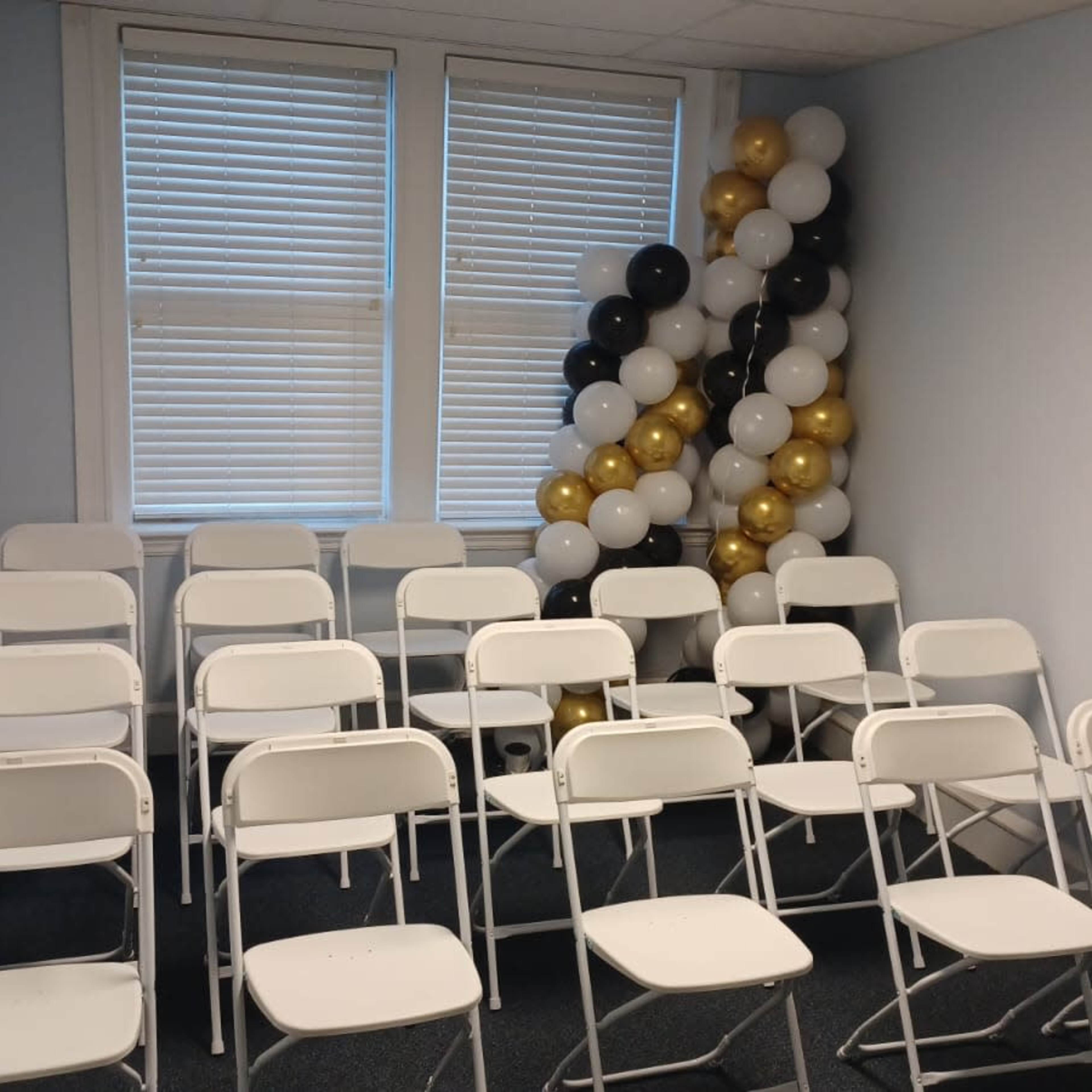 A small room is set up with several white folding chairs arranged in rows, beside a cluster of black, white, and gold balloons.