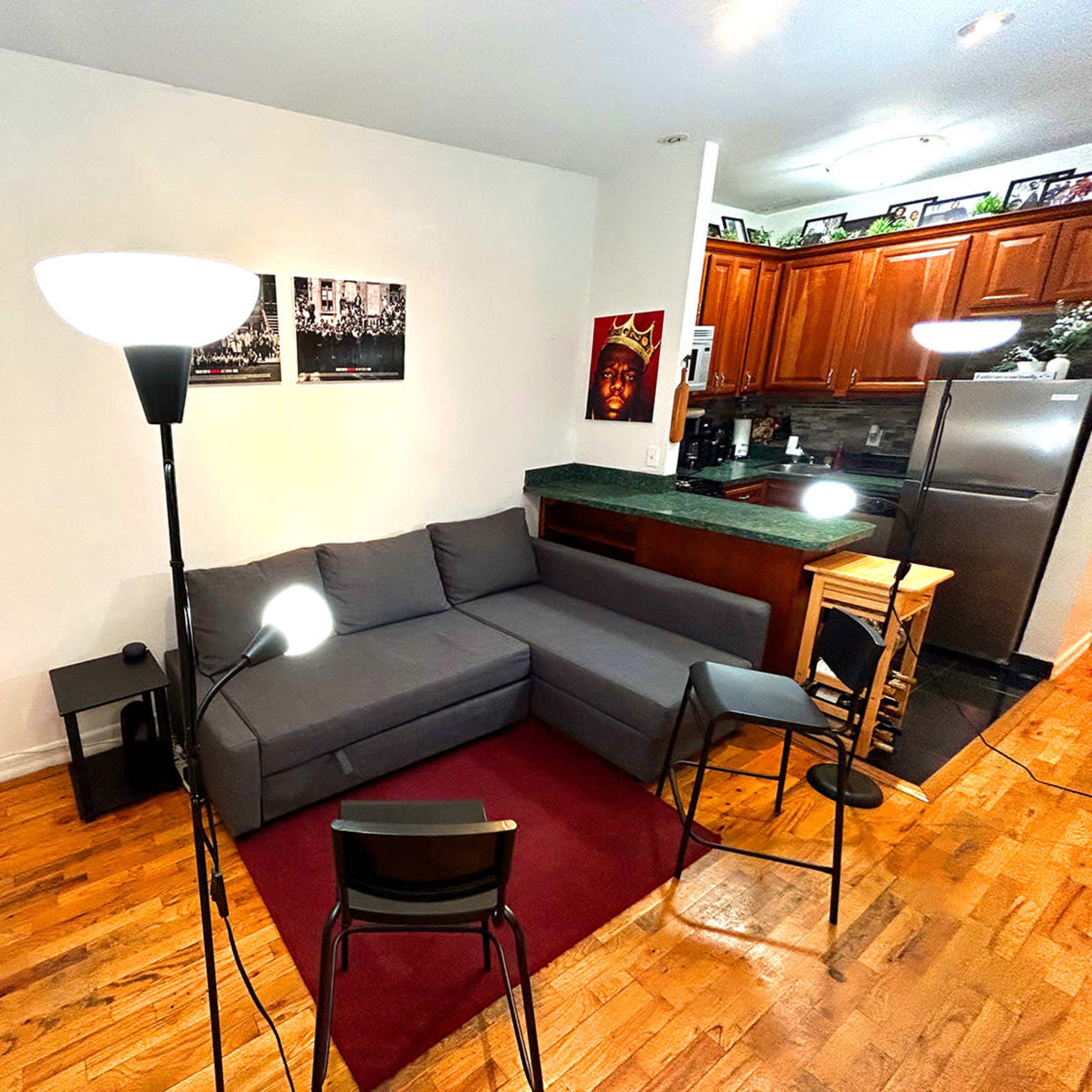 A compact living room with a gray sectional sofa, two floor lamps, a small round table, and a kitchen area in the background.
