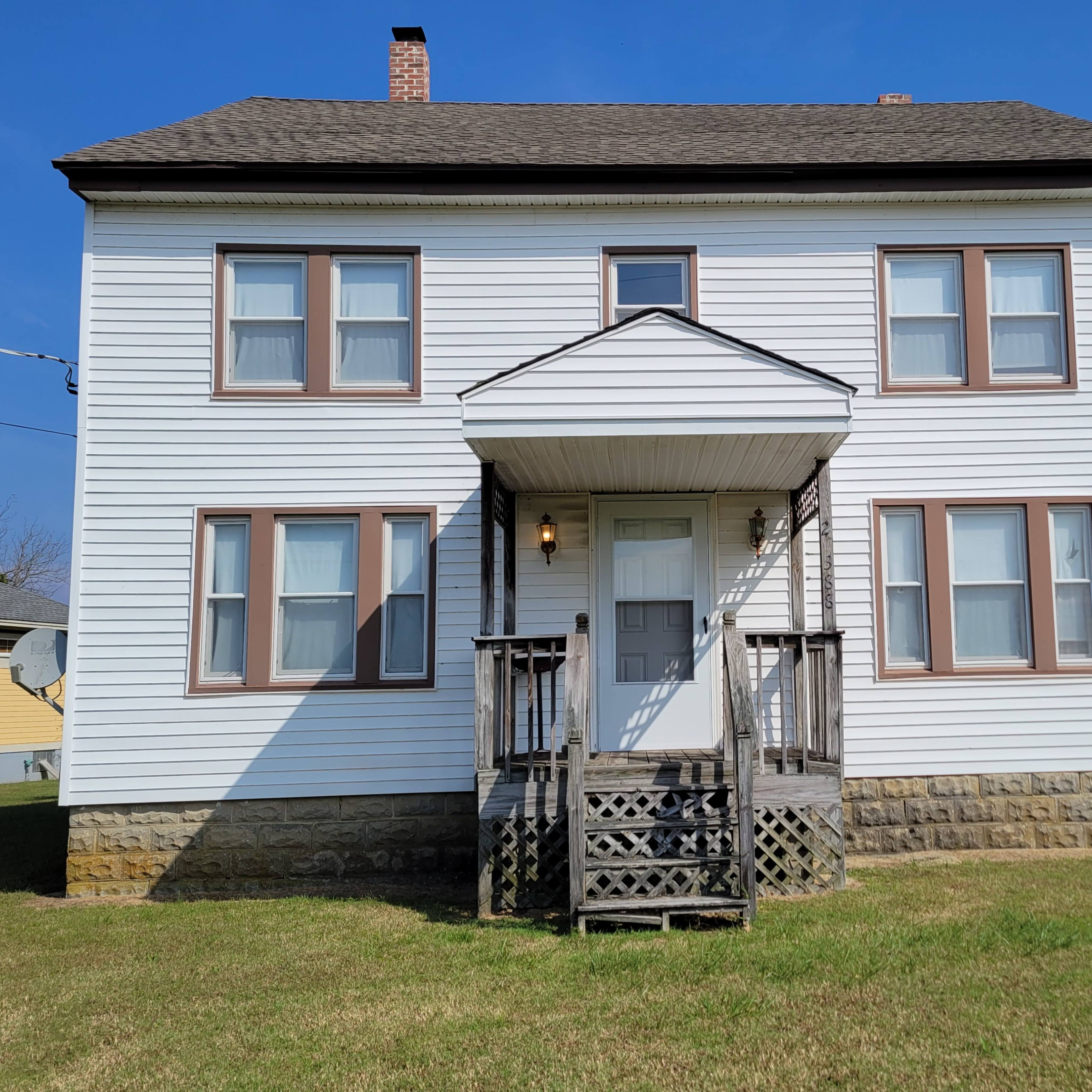 A two-story white house with a staircase and a small porch at the front, surrounded by a grassy yard.