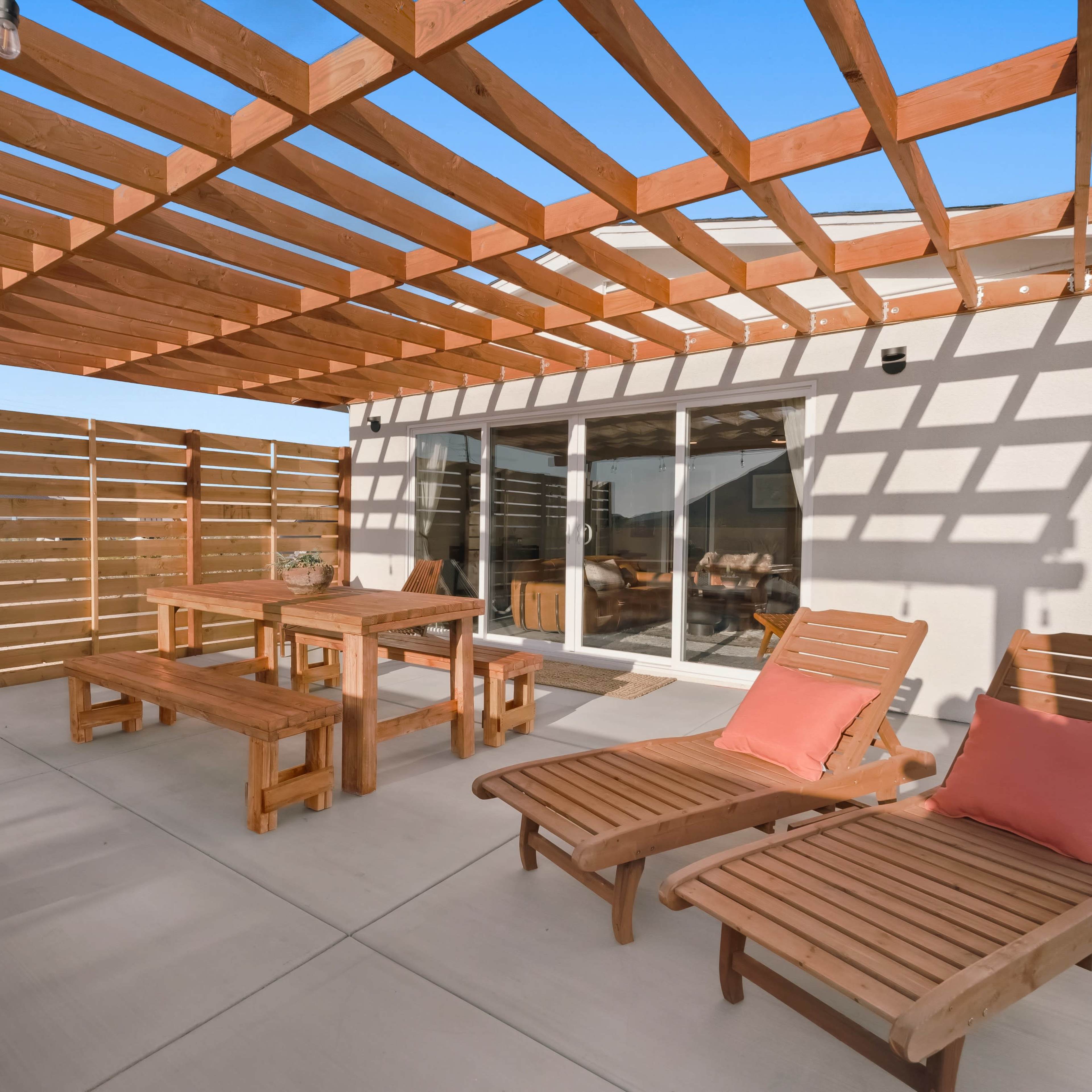 A patio area with wooden furniture, including a table, benches, and lounge chairs, all shaded by a wooden pergola.