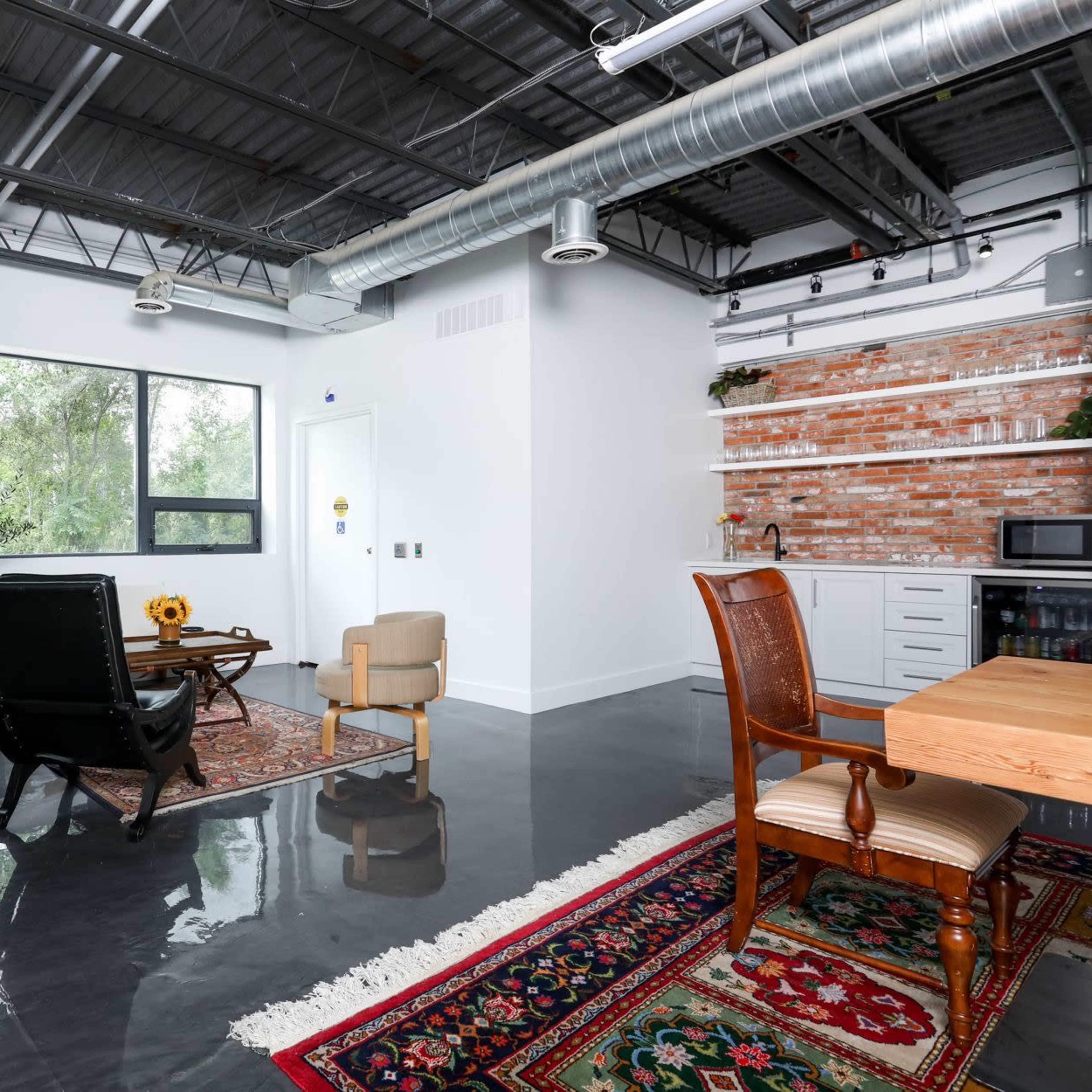 A modern office space featuring a wooden door, a mix of seating options, a dining table, and a kitchen area, with large windows allowing natural light.