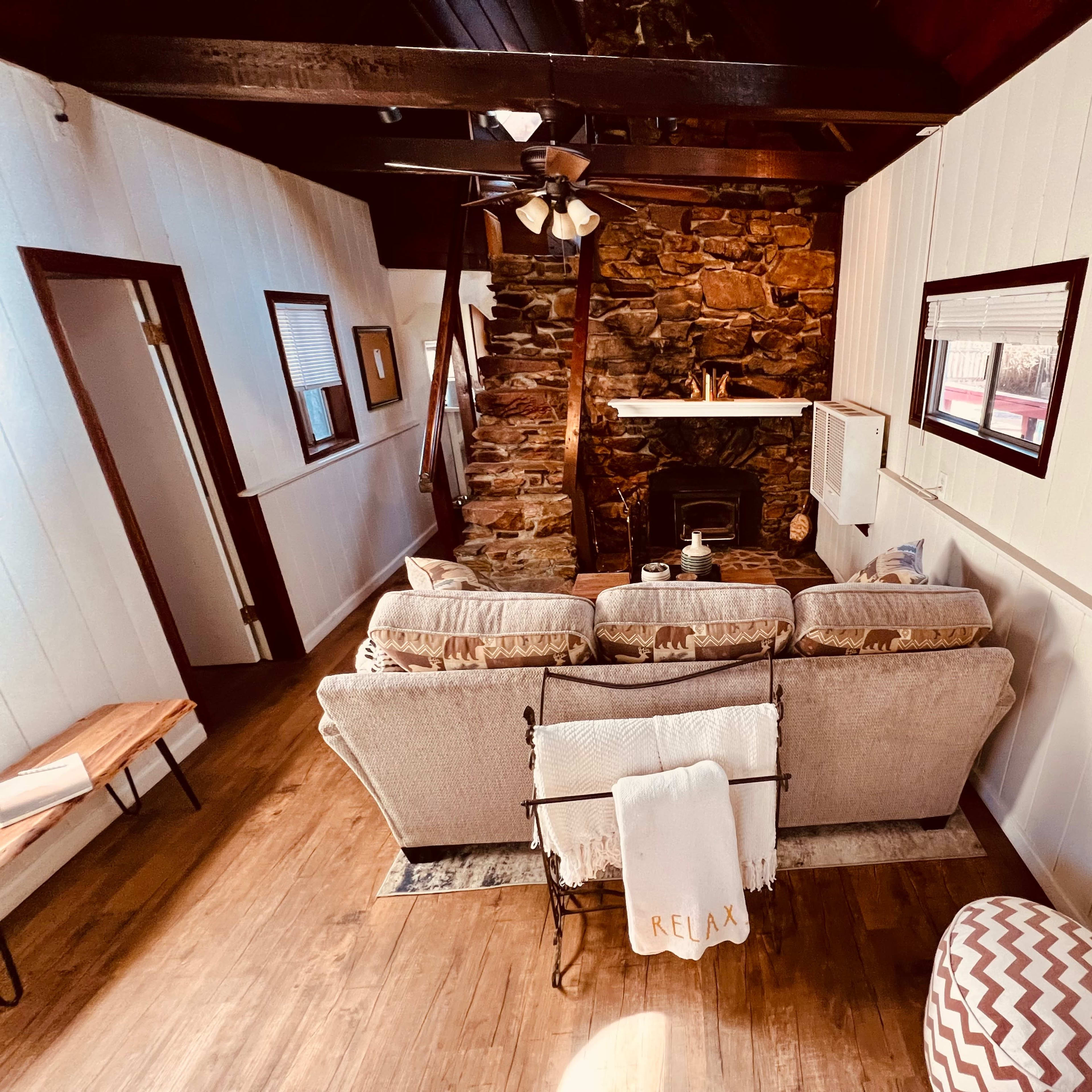 A cozy living room with a stone fireplace, wooden beams, and a seating area featuring a beige couch and a patterned ottoman.