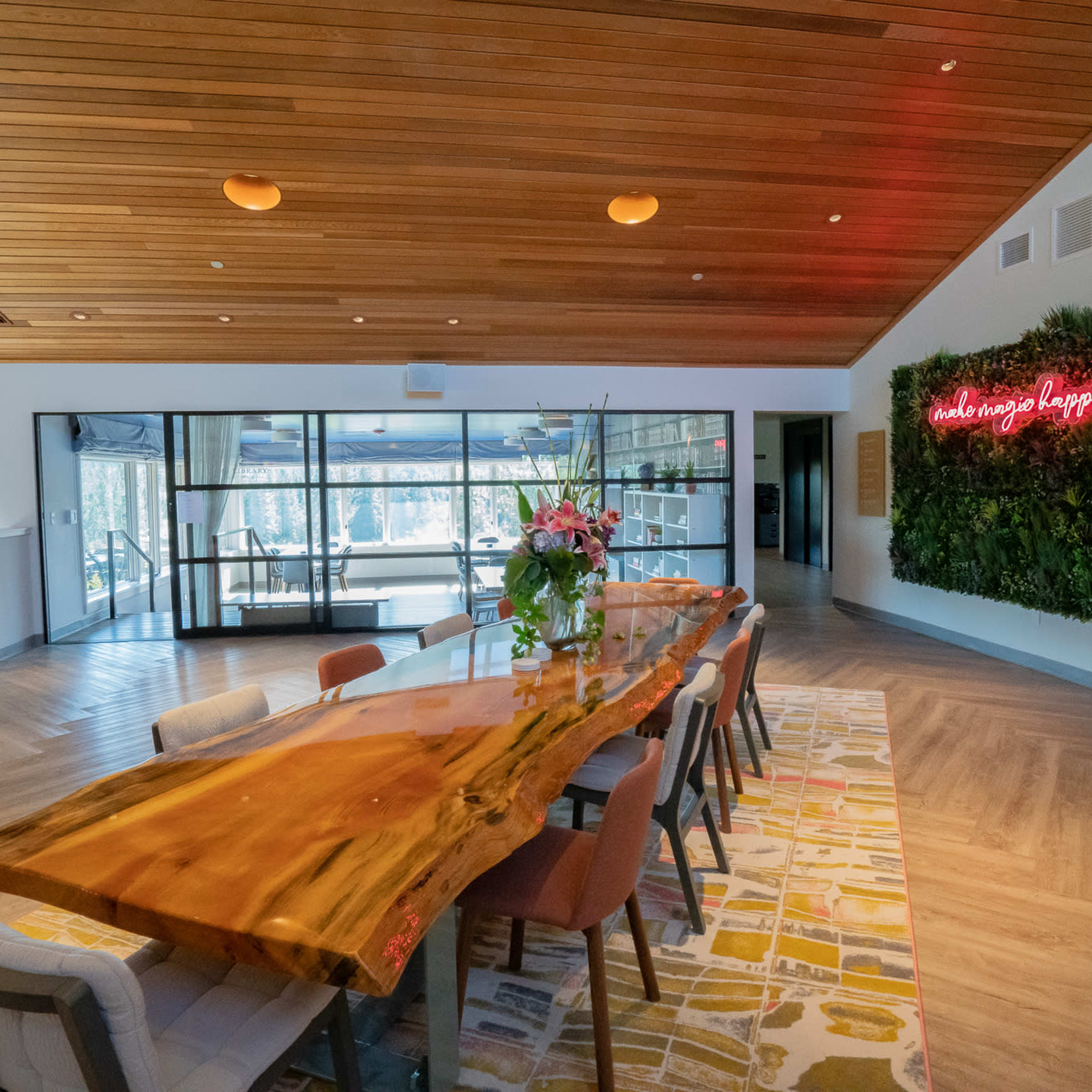 A modern dining area featuring a large wooden table surrounded by chairs, with large windows providing a view of the outdoors and a neon sign on a greenery-covered wall.