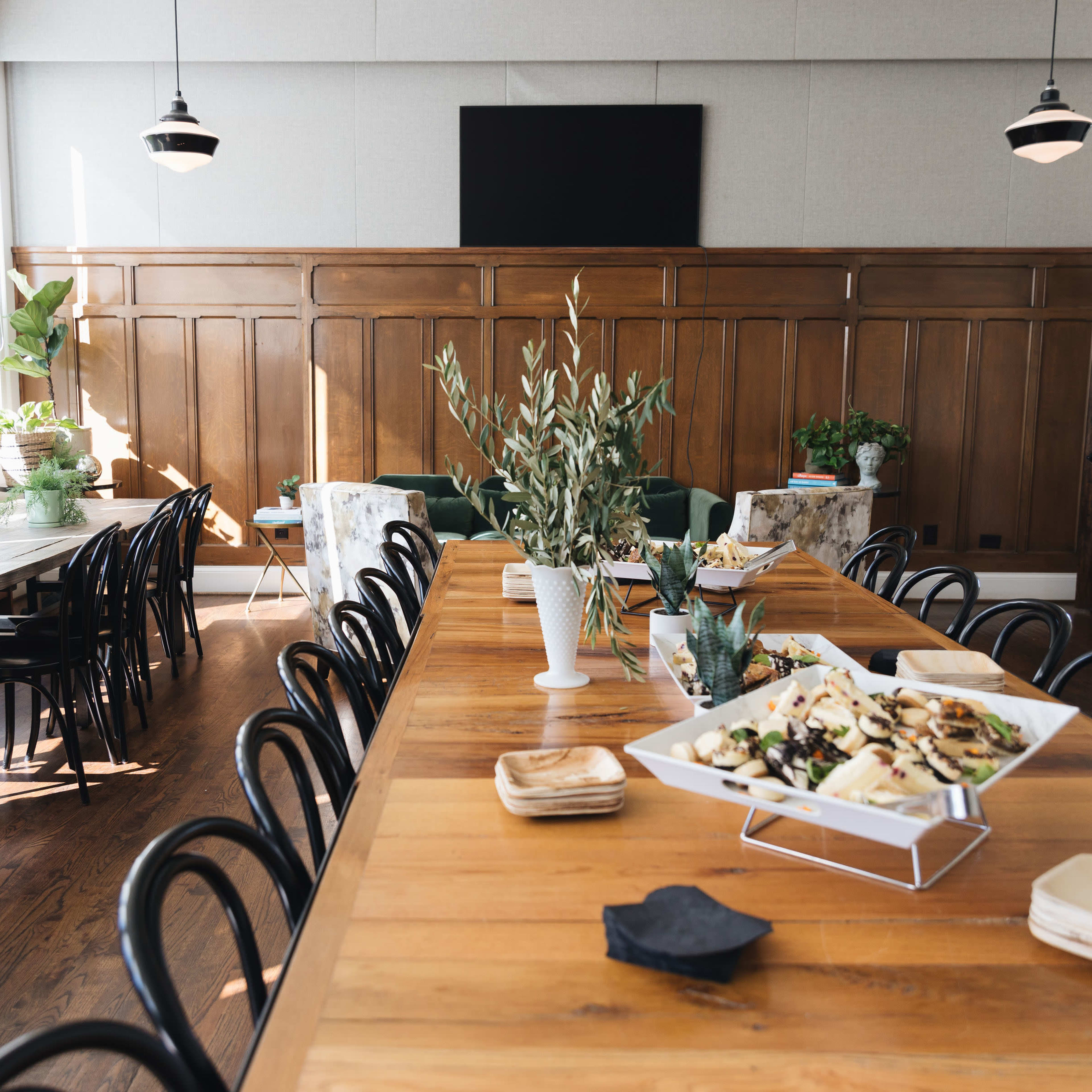A spacious dining room features a long wooden table, decorative plants, and a catering setup with platters of food.