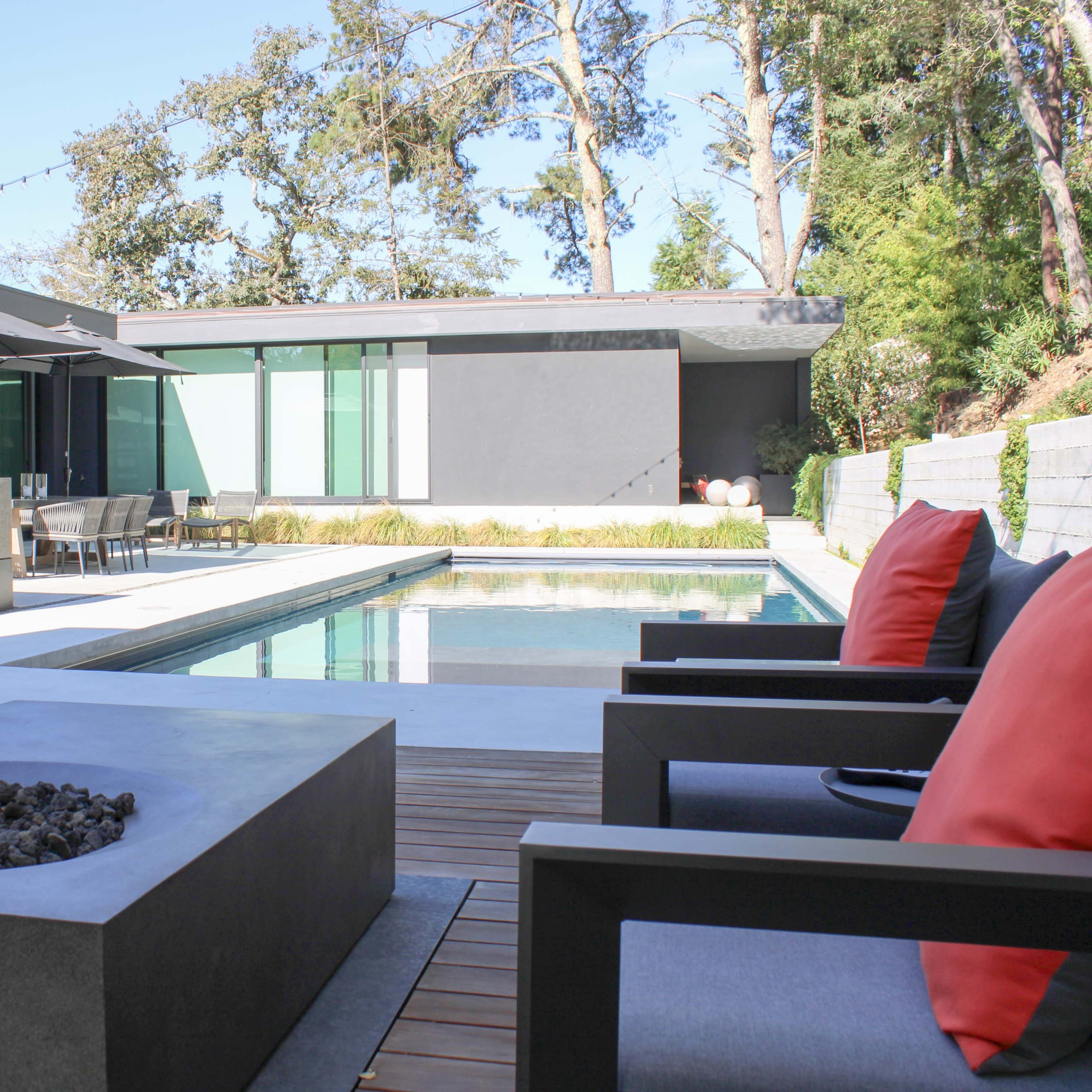 A modern outdoor space featuring a swimming pool, a fire pit, and patio furniture surrounded by greenery.