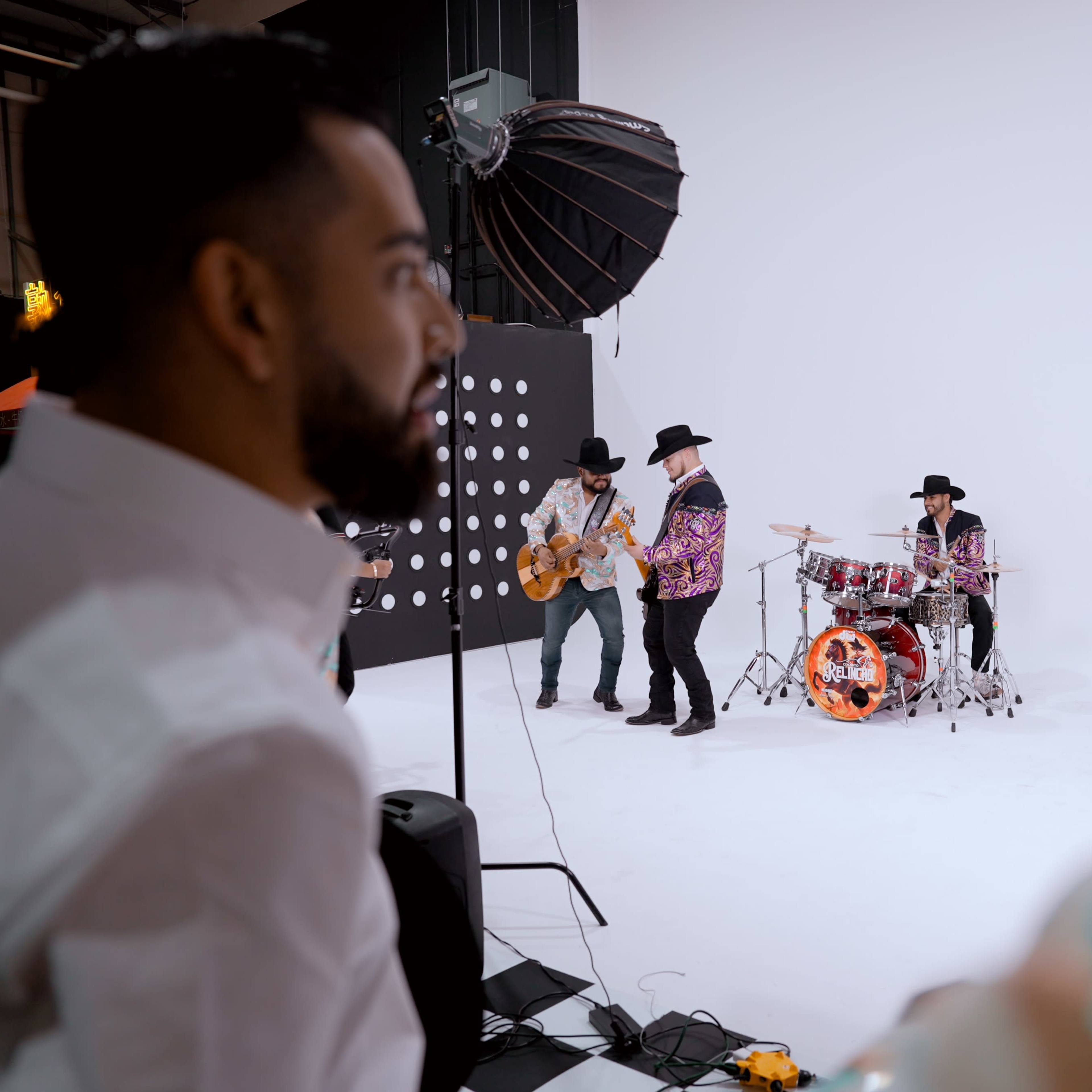 Two men watch a band performing with instruments against a plain white backdrop in a studio setting.