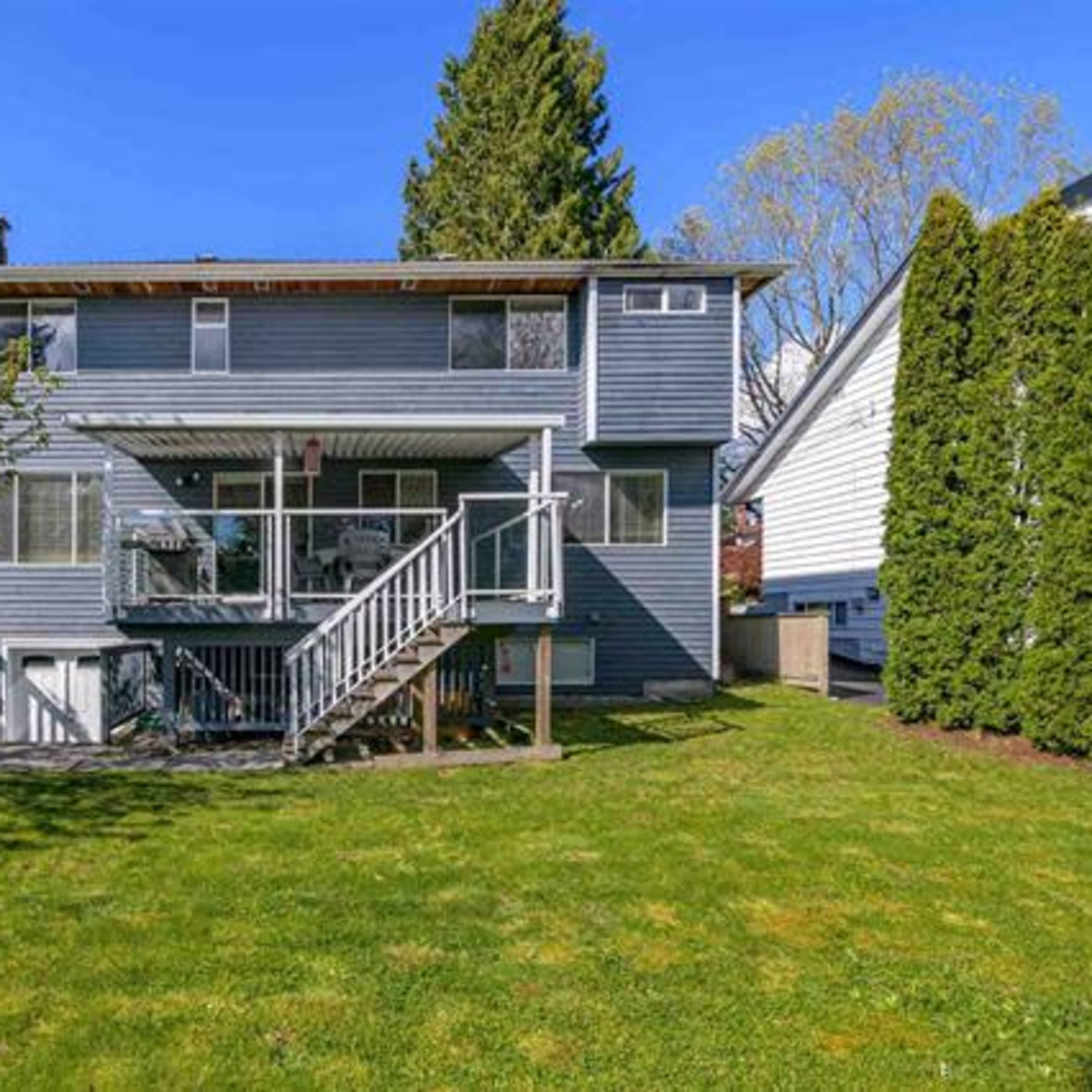 The back exterior of a two-story house with a deck and a well-maintained grassy yard, surrounded by trees and neighboring homes.