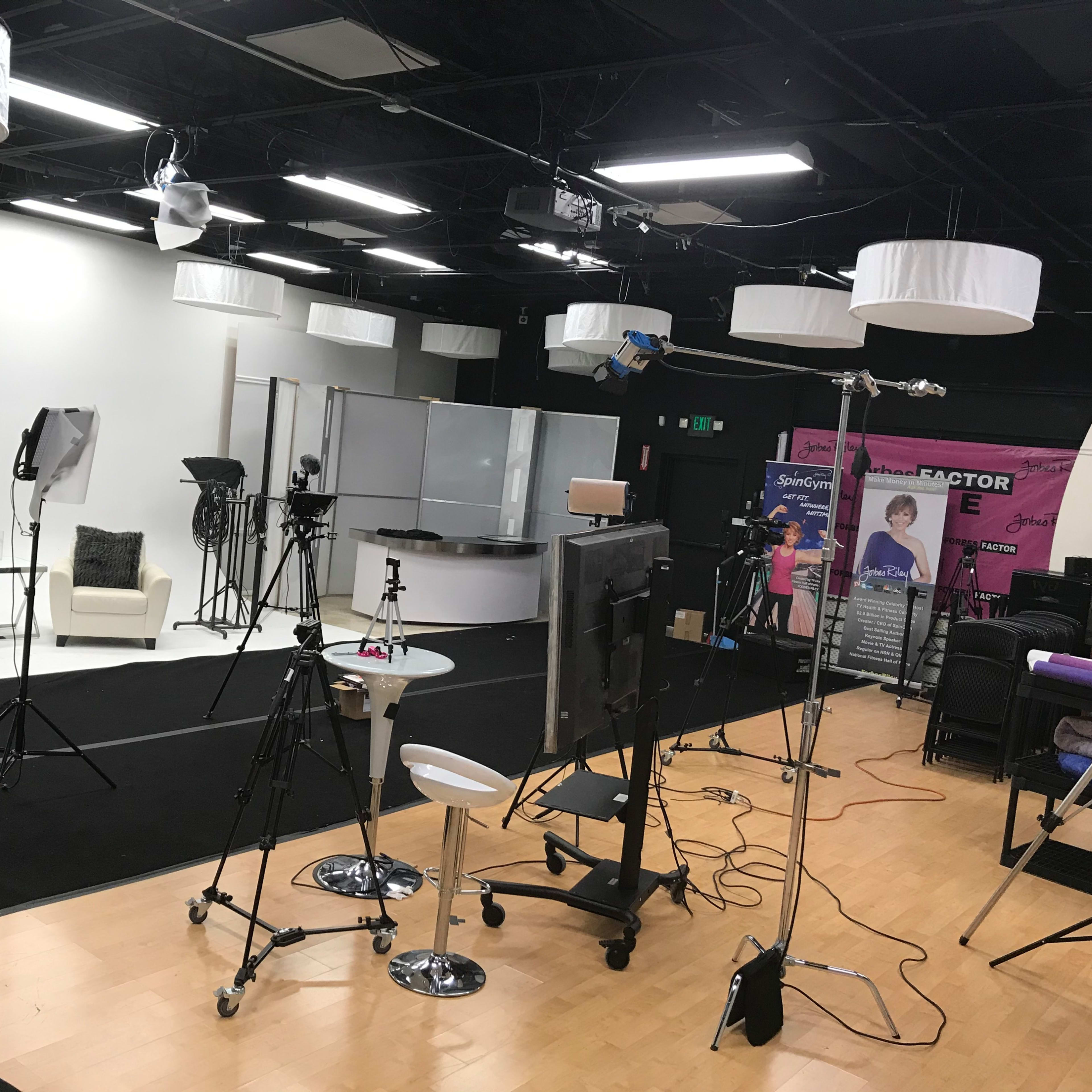 A studio set up for a production, featuring a seating area, multiple cameras, and lighting equipment arranged on a black carpeted floor.