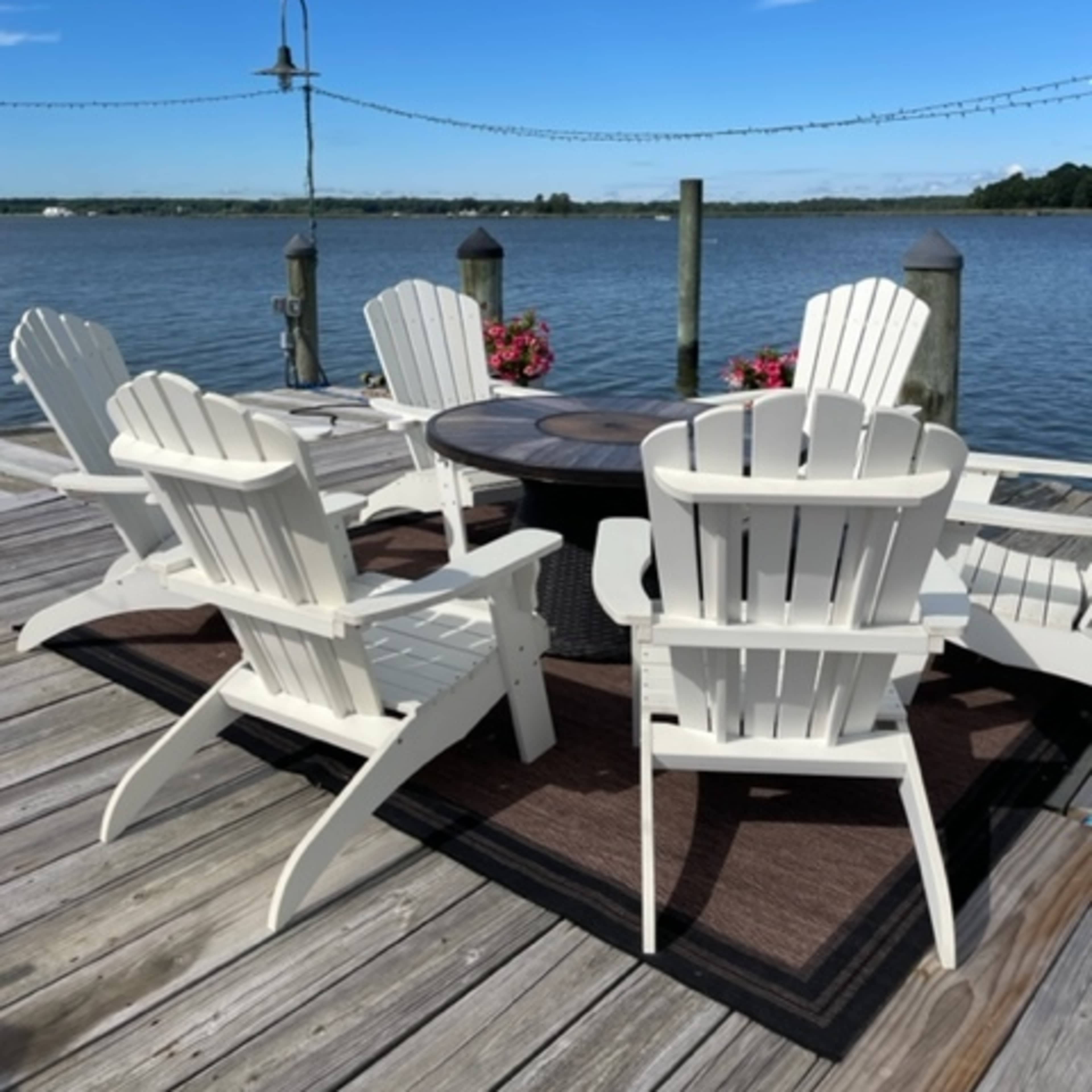 A set of white Adirondack chairs surrounds a circular table on a wooden dock overlooking the water.