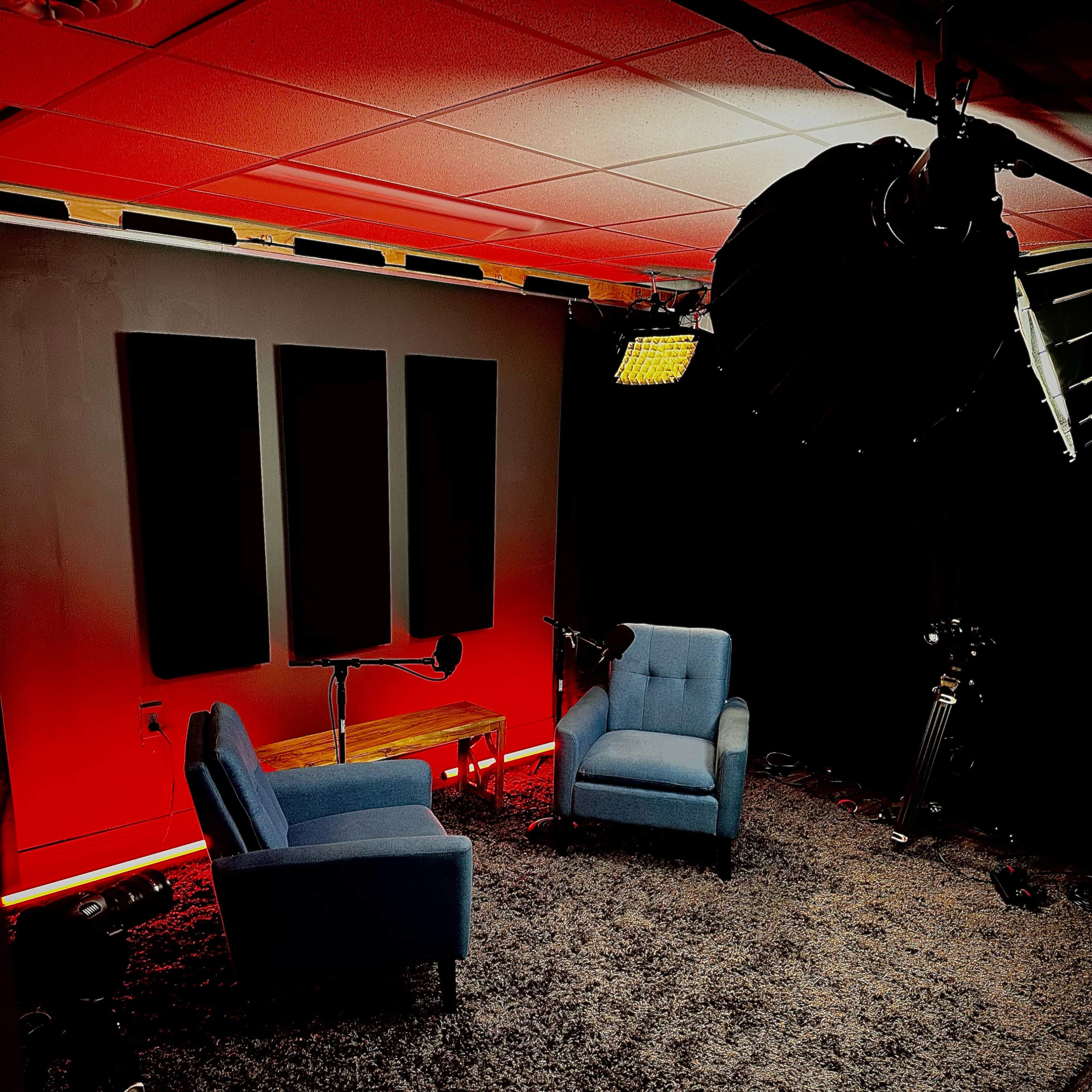 A small, dimly lit studio setup featuring two blue armchairs facing each other on a carpeted floor, with soft lighting and microphones positioned nearby.