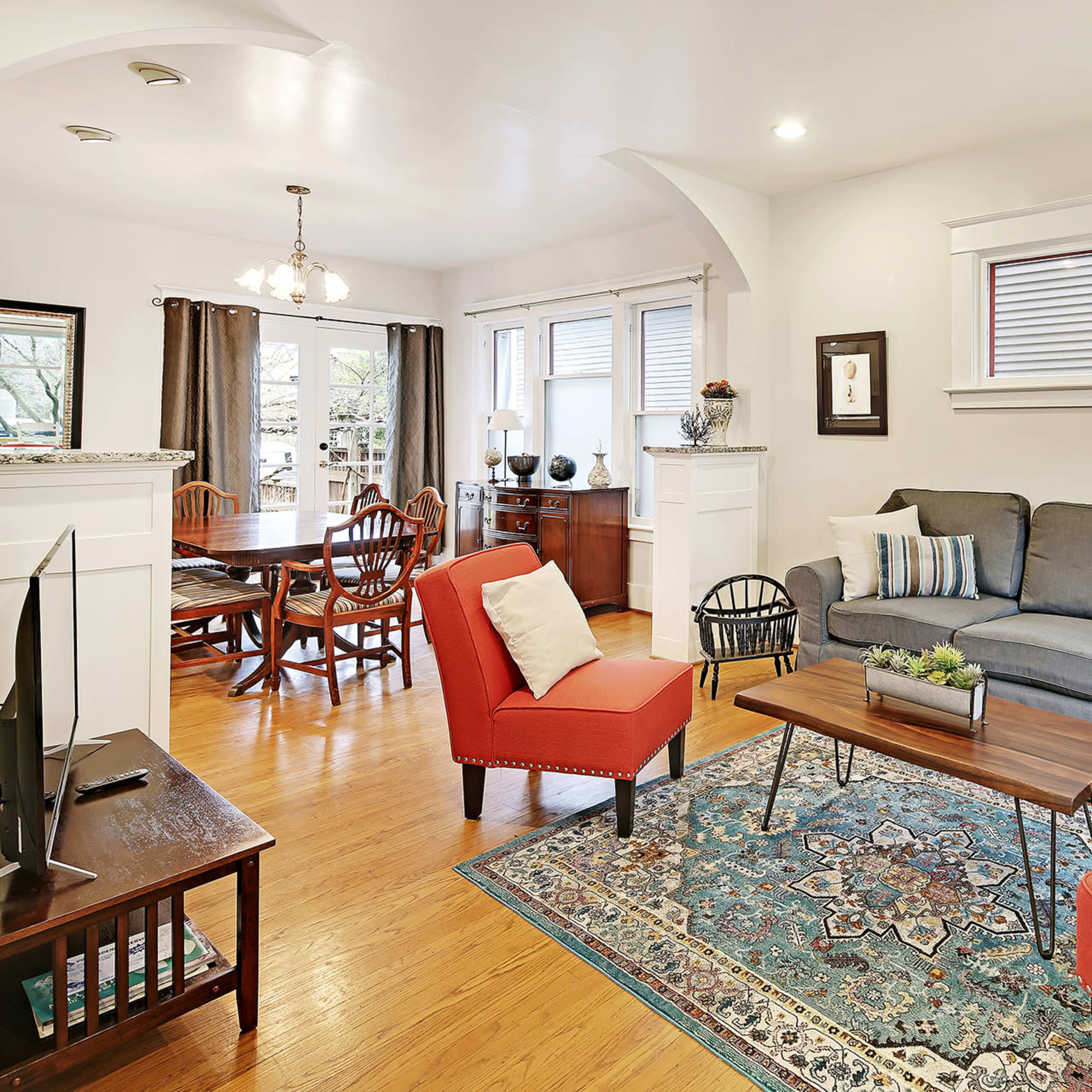 A living room with a gray couch, two red chairs, a wooden coffee table on a decorative rug, and a dining area in the background.