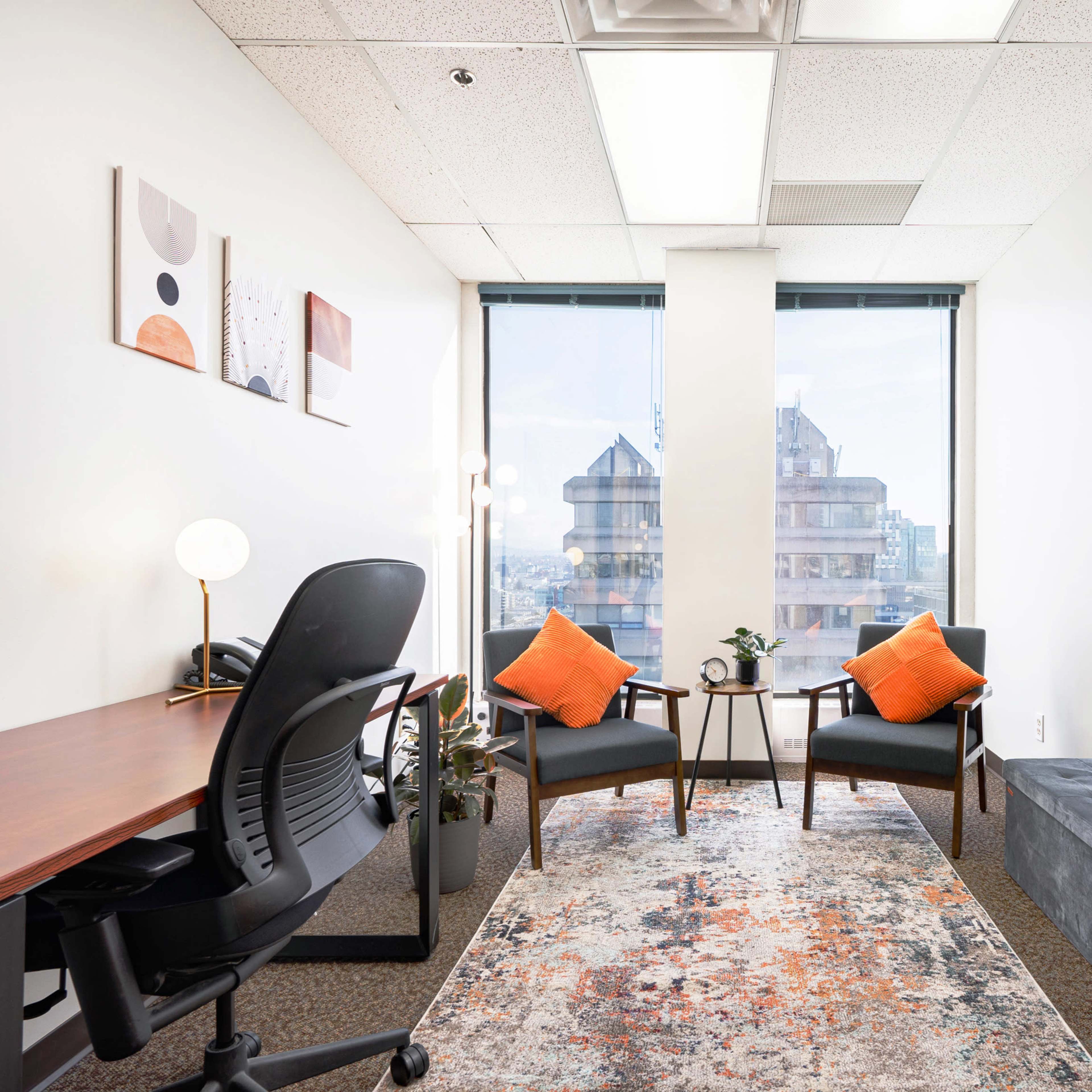 A small office with two chairs and a table, featuring a desk and a window that overlooks a cityscape.