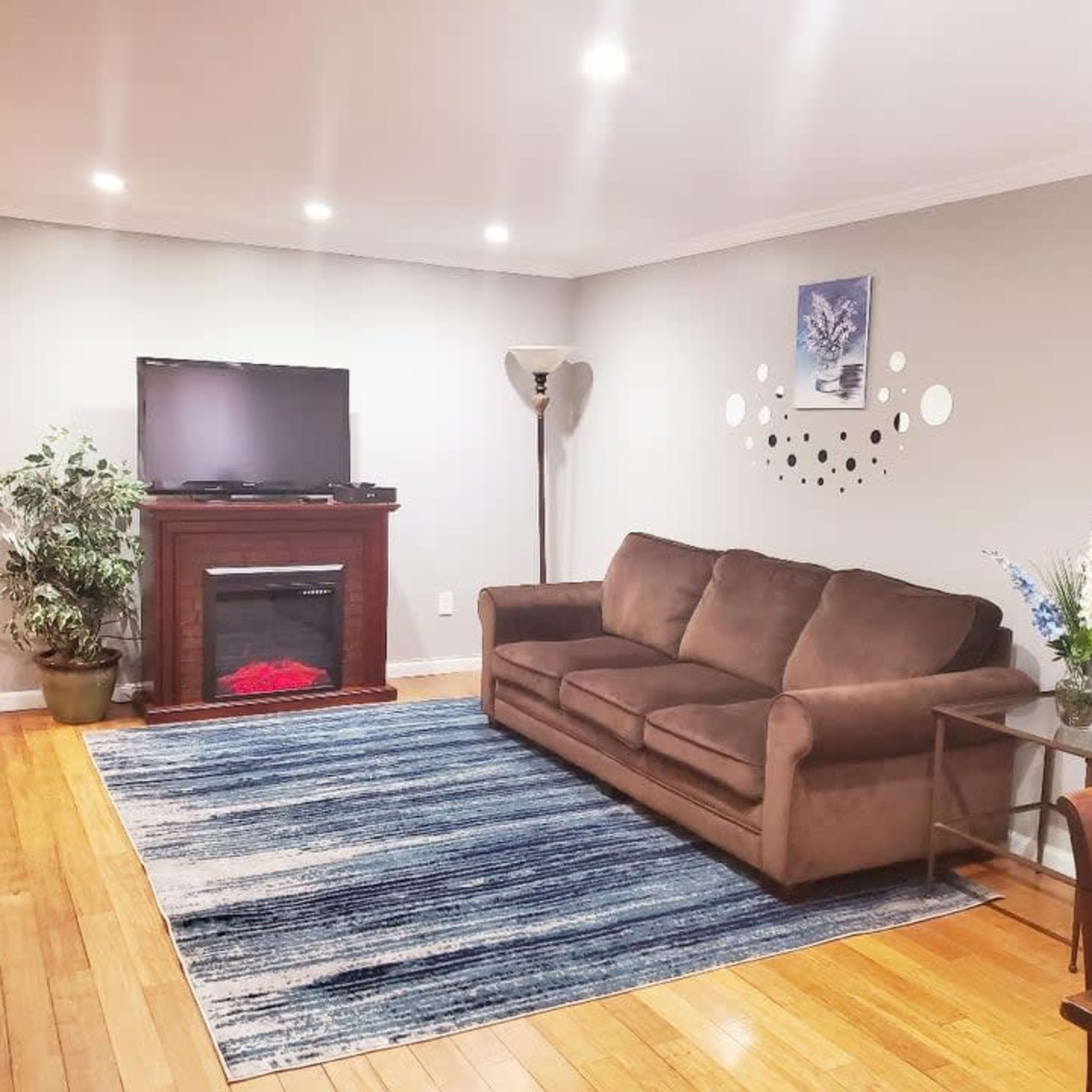 A living room featuring a brown sofa, a television on a wooden stand with an electric fireplace below, a patterned area rug, a small table, and a vase with flowers.
