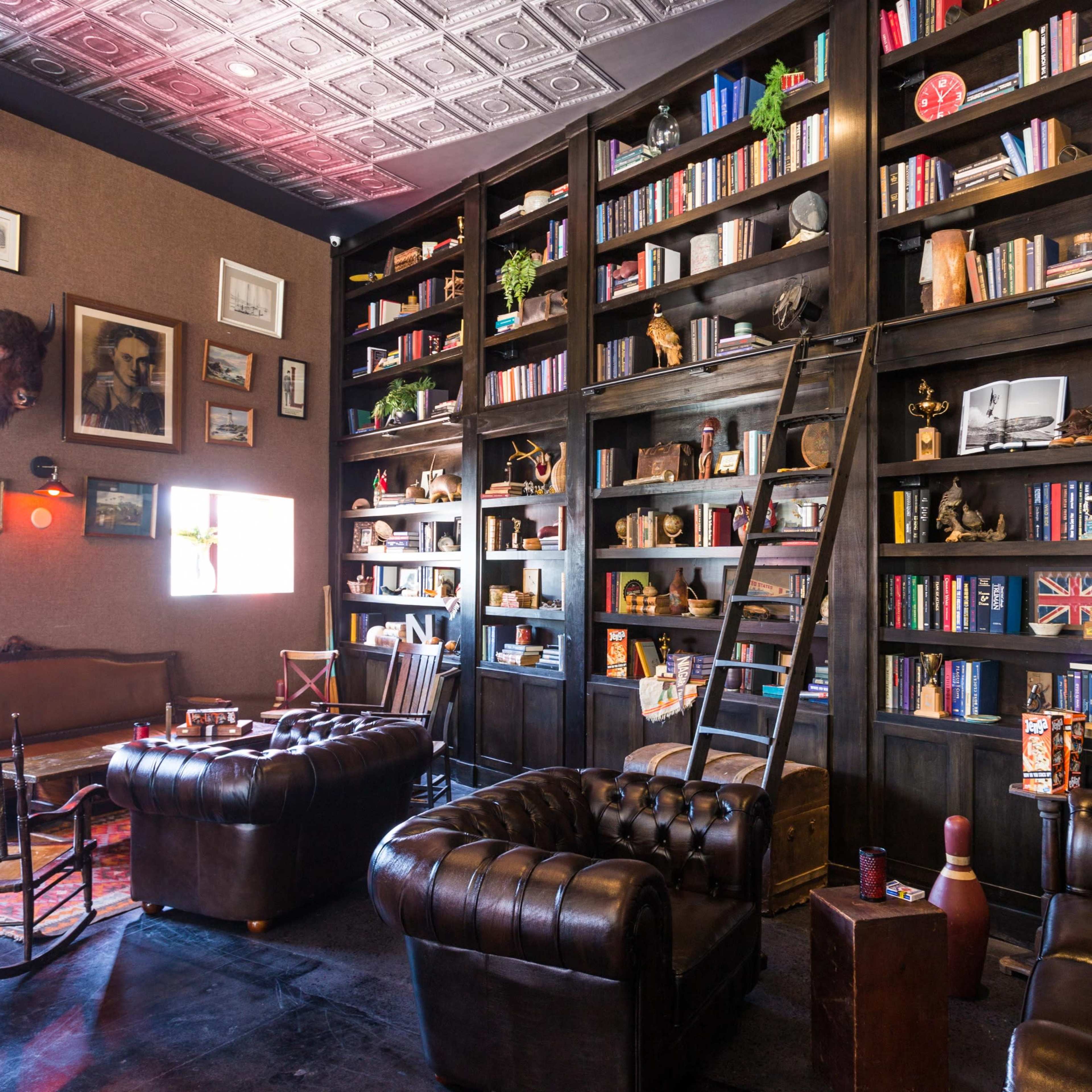 A cozy reading room featuring a large black bookshelf filled with books and decorative items, accompanied by leather seating and a wooden ladder.