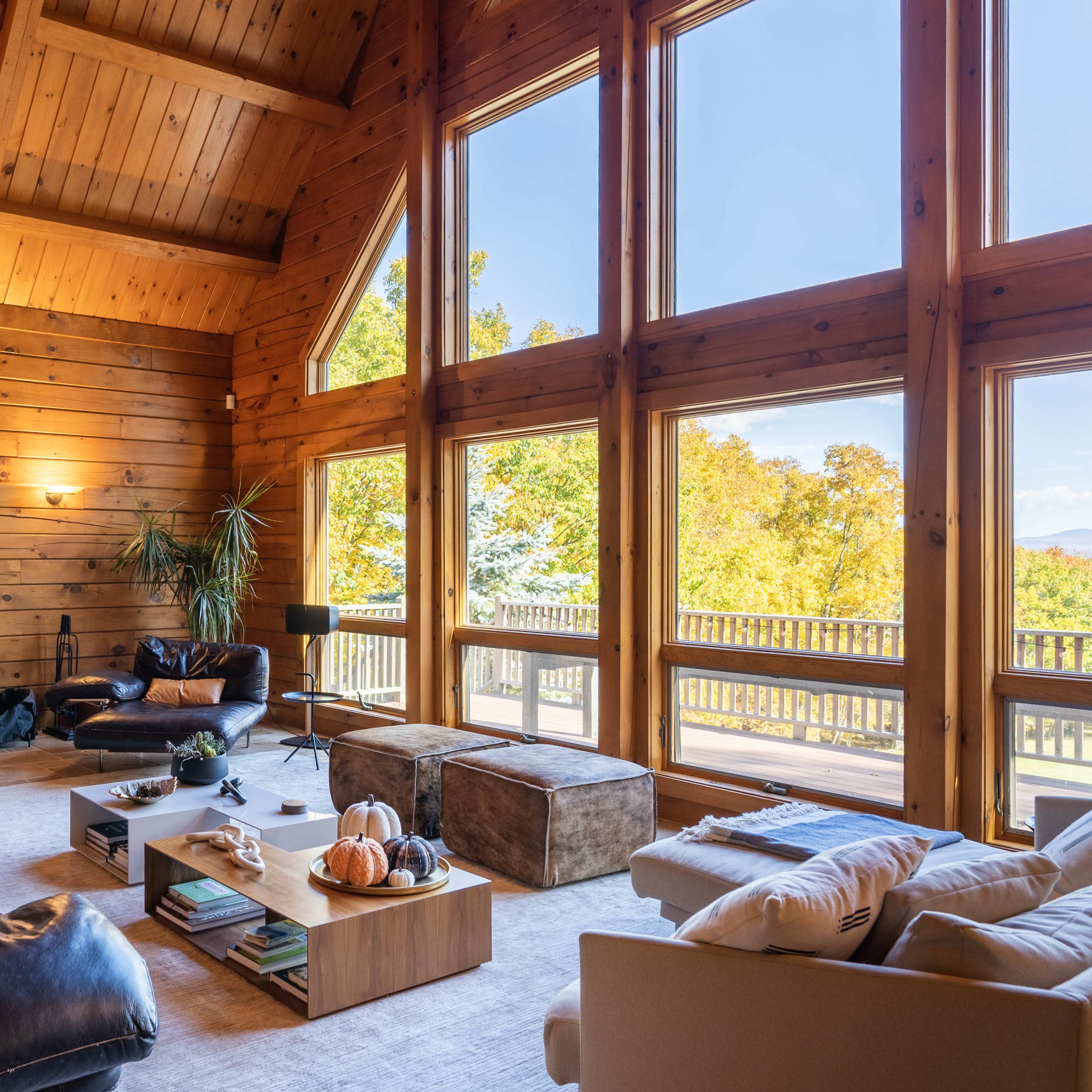 The interior of a wooden cabin features a spacious living area with large windows overlooking trees and a fireplace at one end.