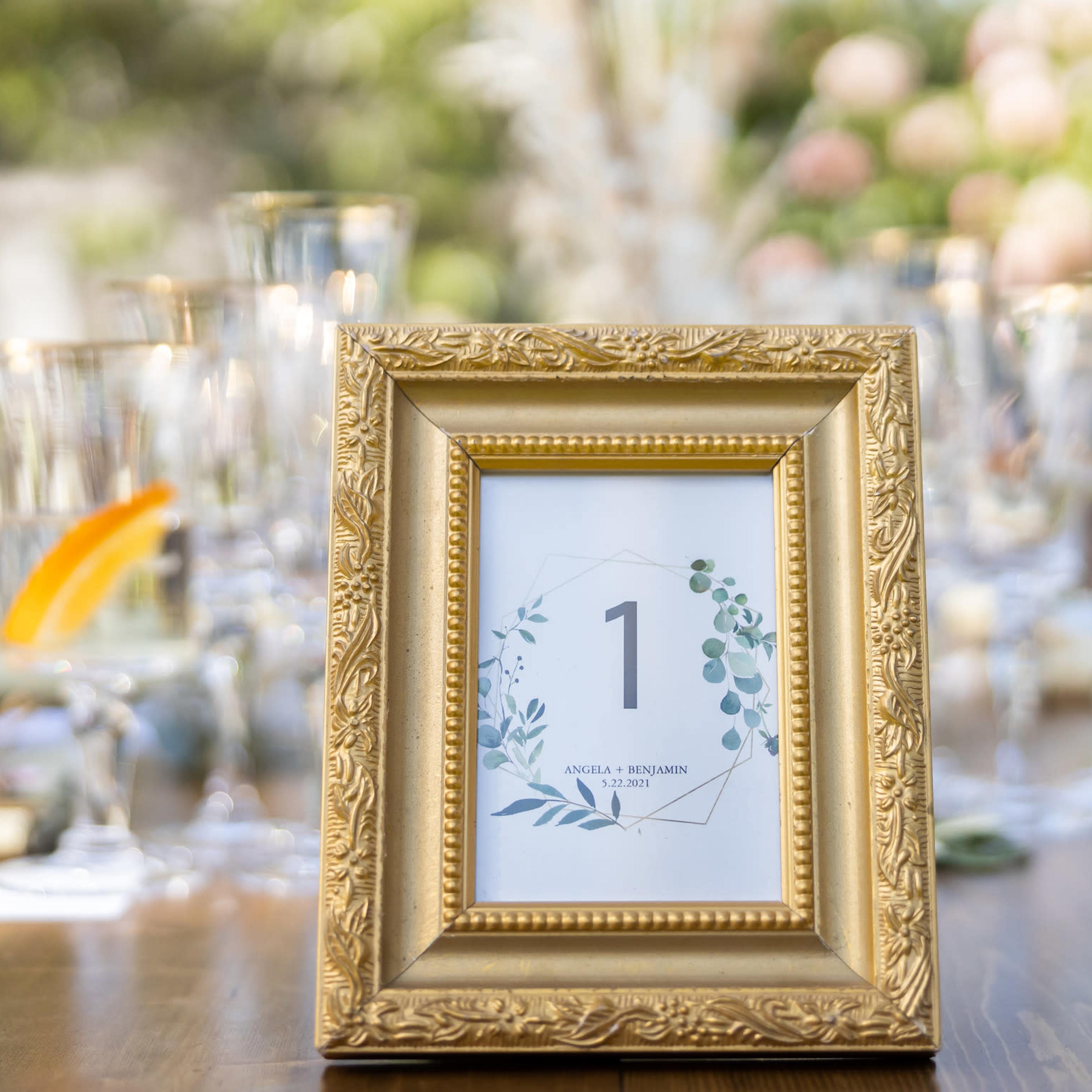A gold-framed table number display is set on a wooden table, surrounded by glassware and decorative elements, indicating the number one.
