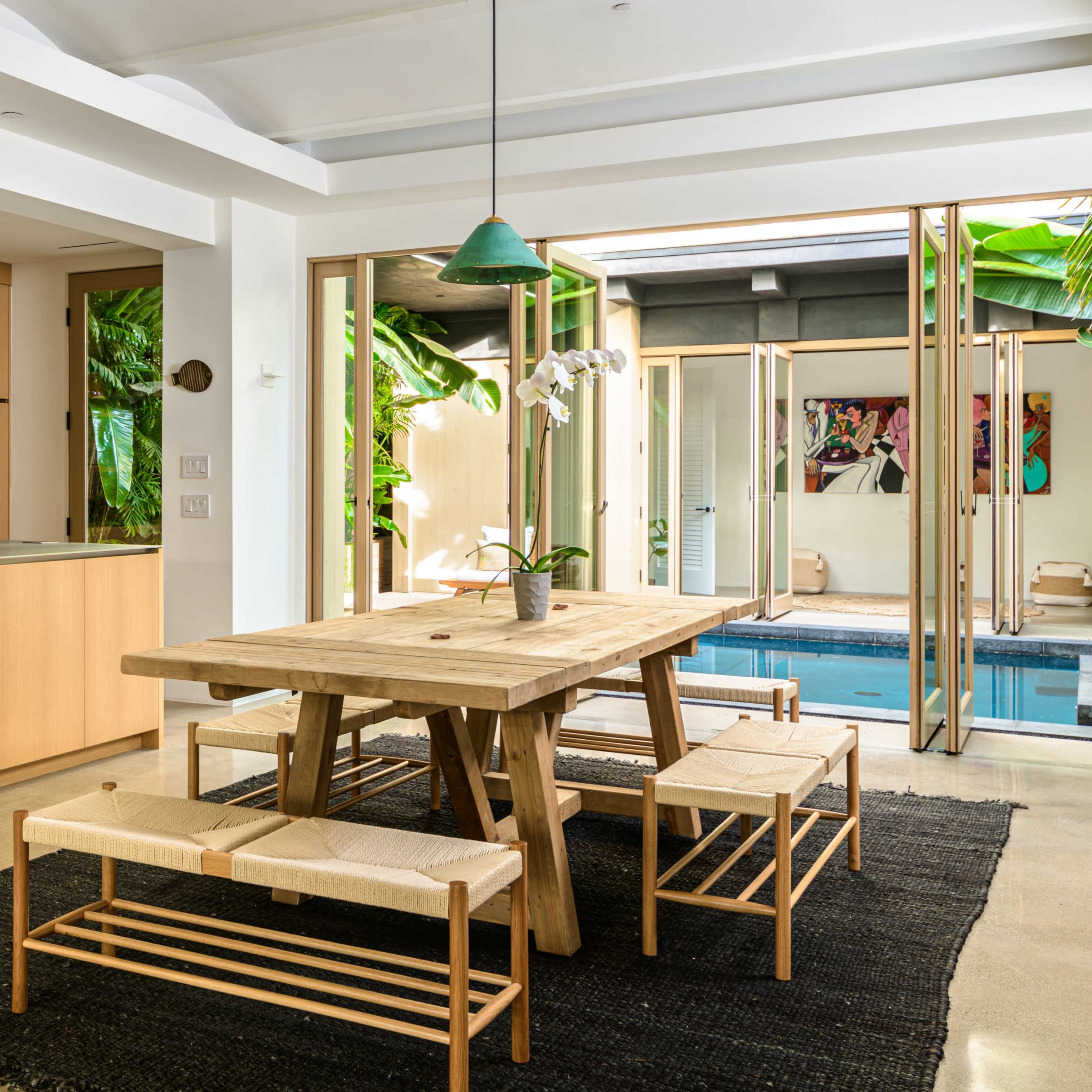 A modern dining area with a wooden table and benches, surrounded by large glass doors that open to a pool area and lush greenery.