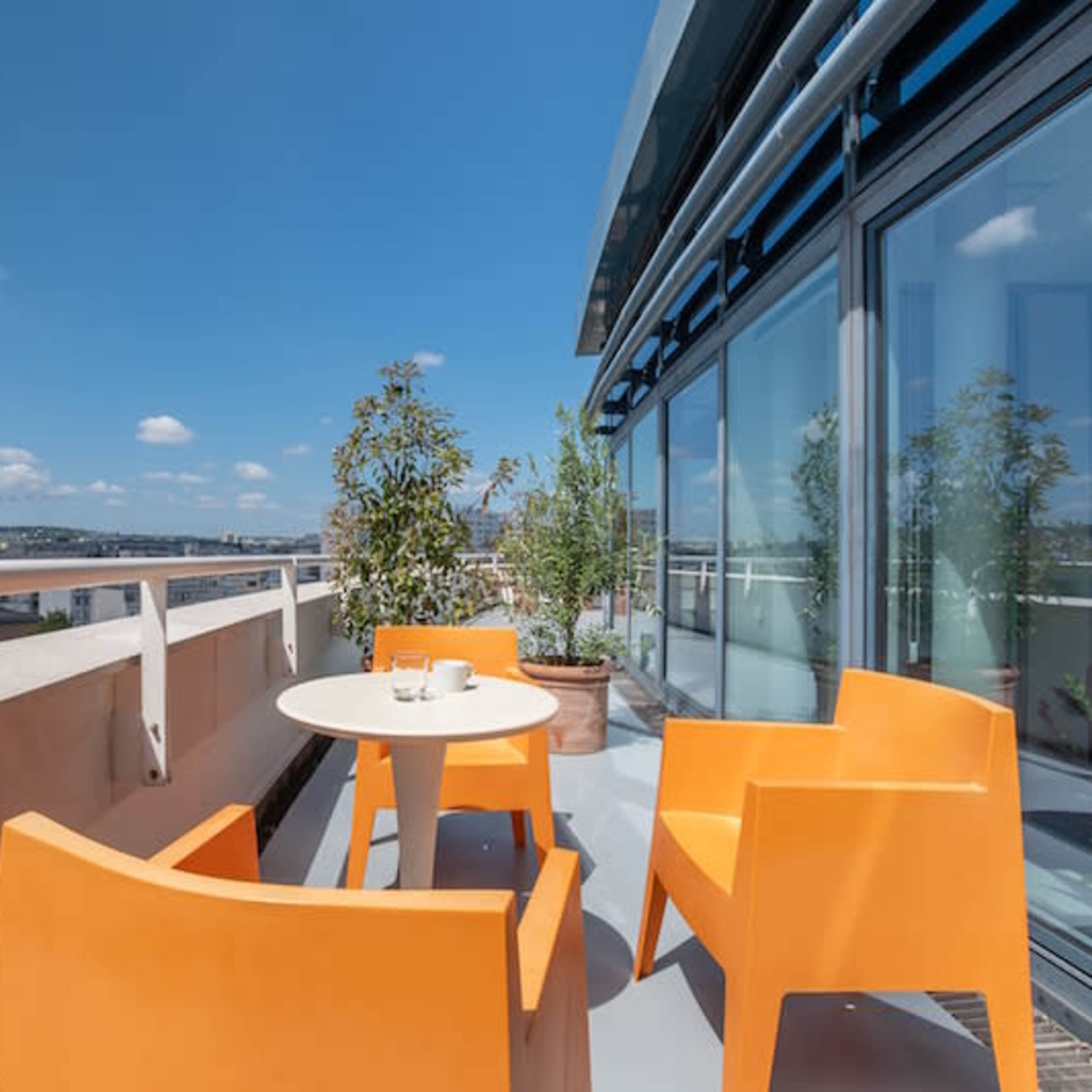 A rooftop terrace with orange chairs and a small table, surrounded by potted plants and a clear blue sky.