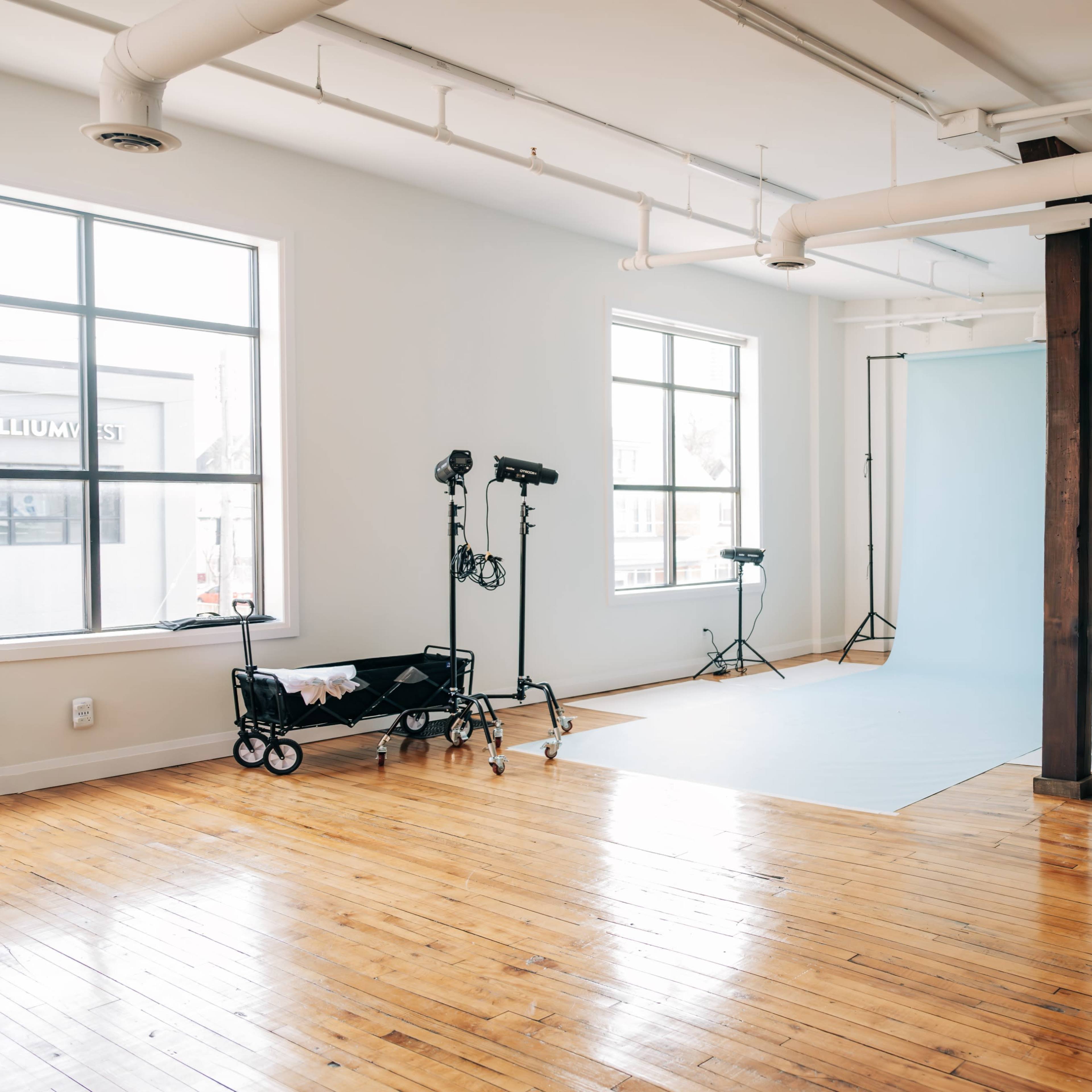 A spacious photography studio with large windows, wooden floors, and backdrops set up on stands.