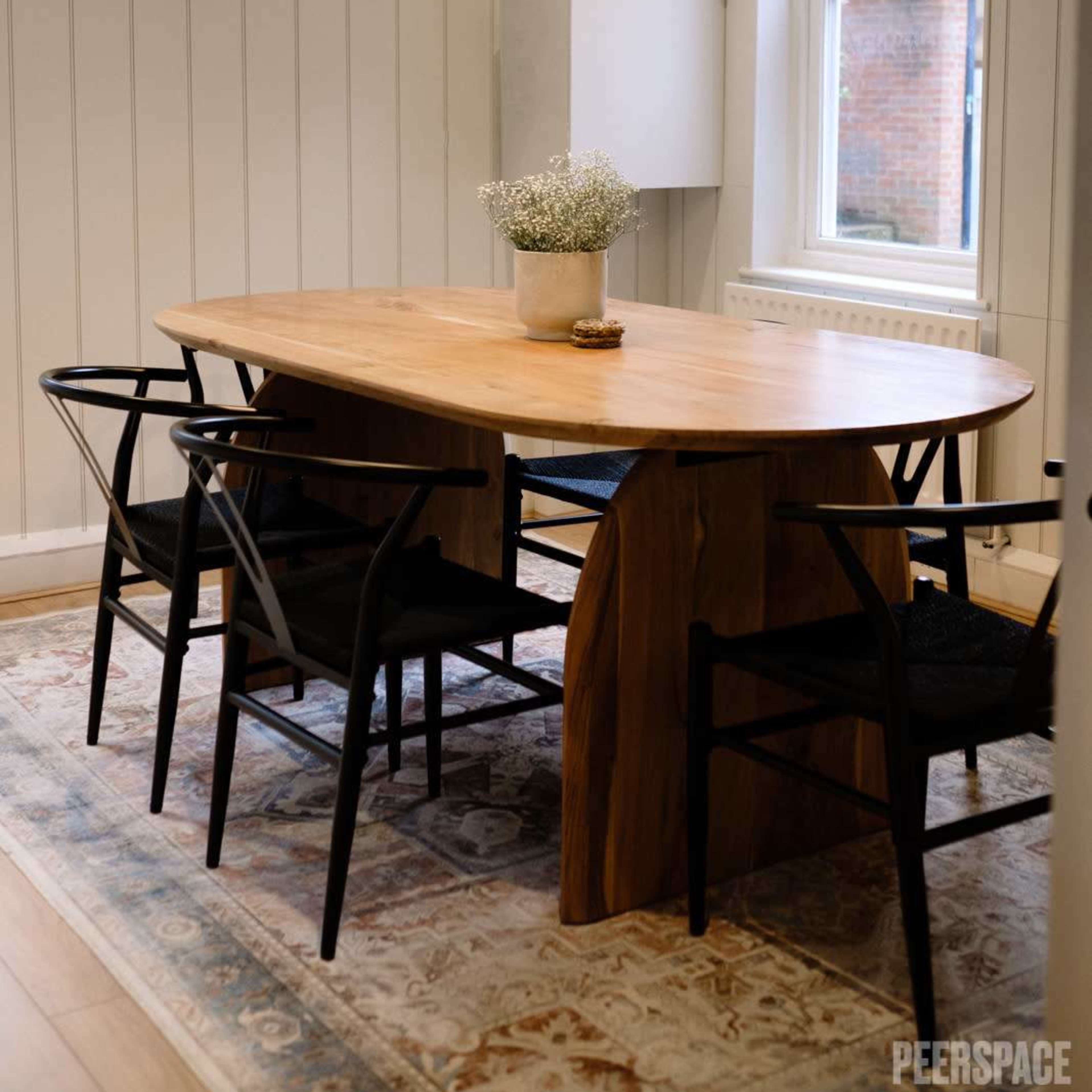 A light wood dining table with black chairs is centered under three pendant lights in a room with white paneled walls and a window.