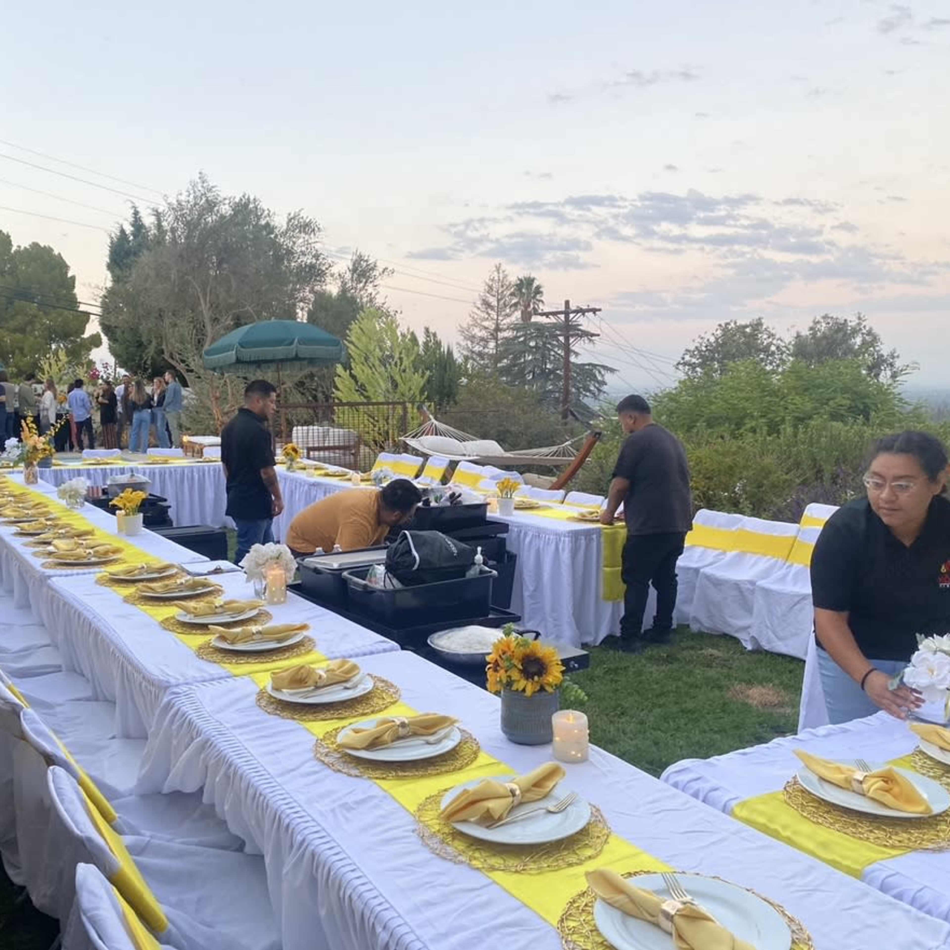 A long outdoor dining setup is prepared with white tablecloths, yellow accents, and floral centerpieces, while staff members arrange the tables before an event.