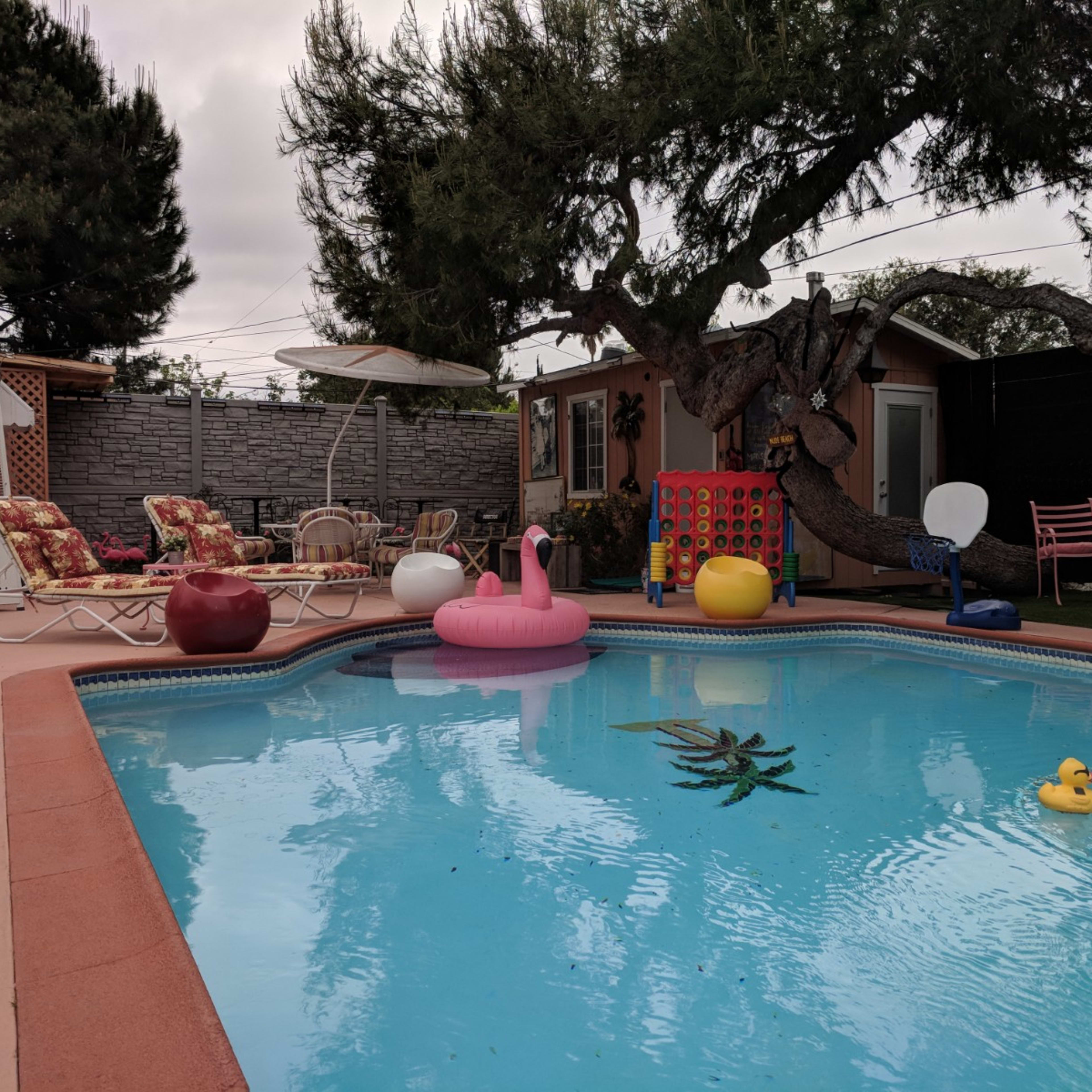 A swimming pool is surrounded by colorful lounge chairs and inflatable decorations, with a backdrop of a house and trees.