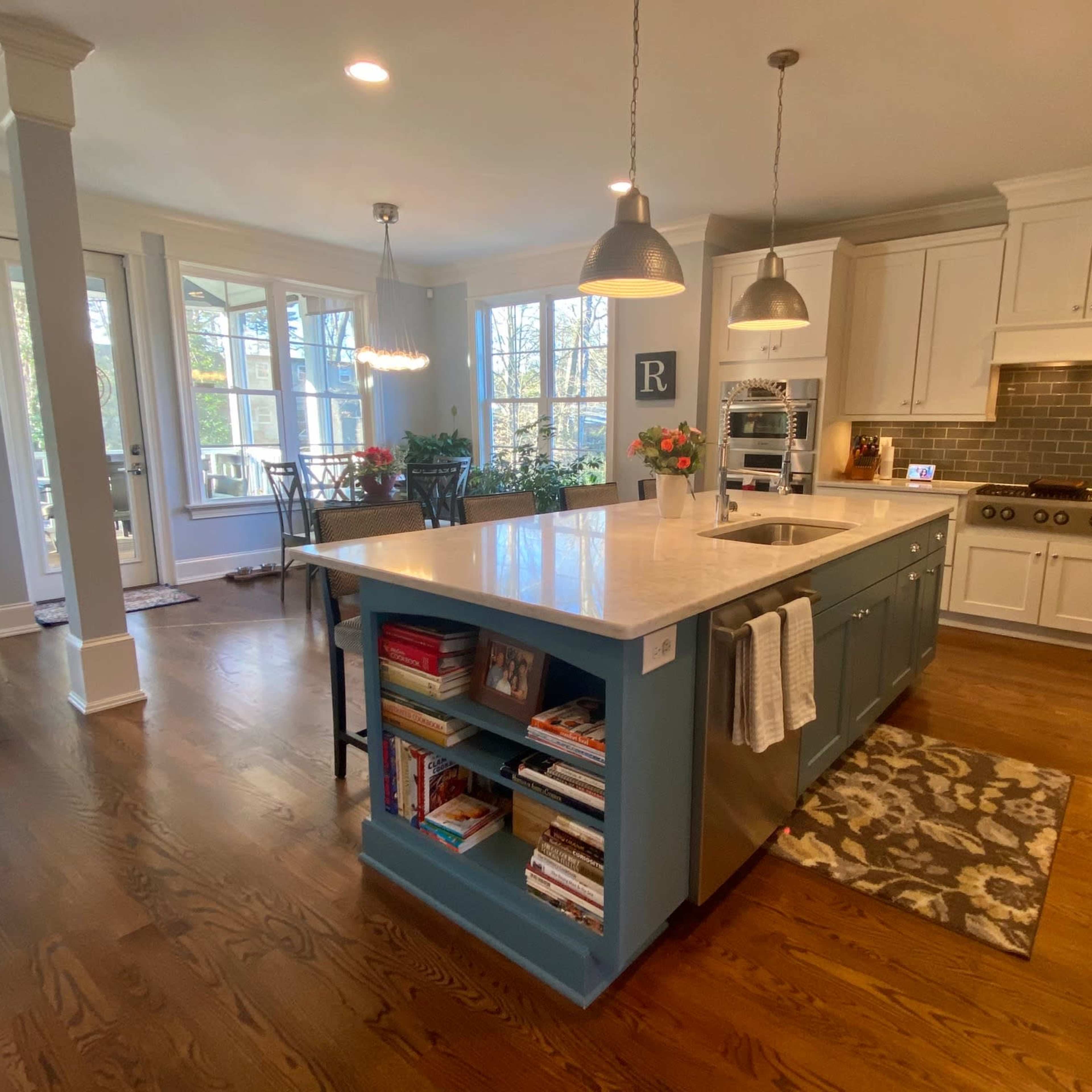 A modern kitchen featuring a large central island with blue cabinetry, white countertops, and stainless steel appliances, illuminated by overhead pendant lights.