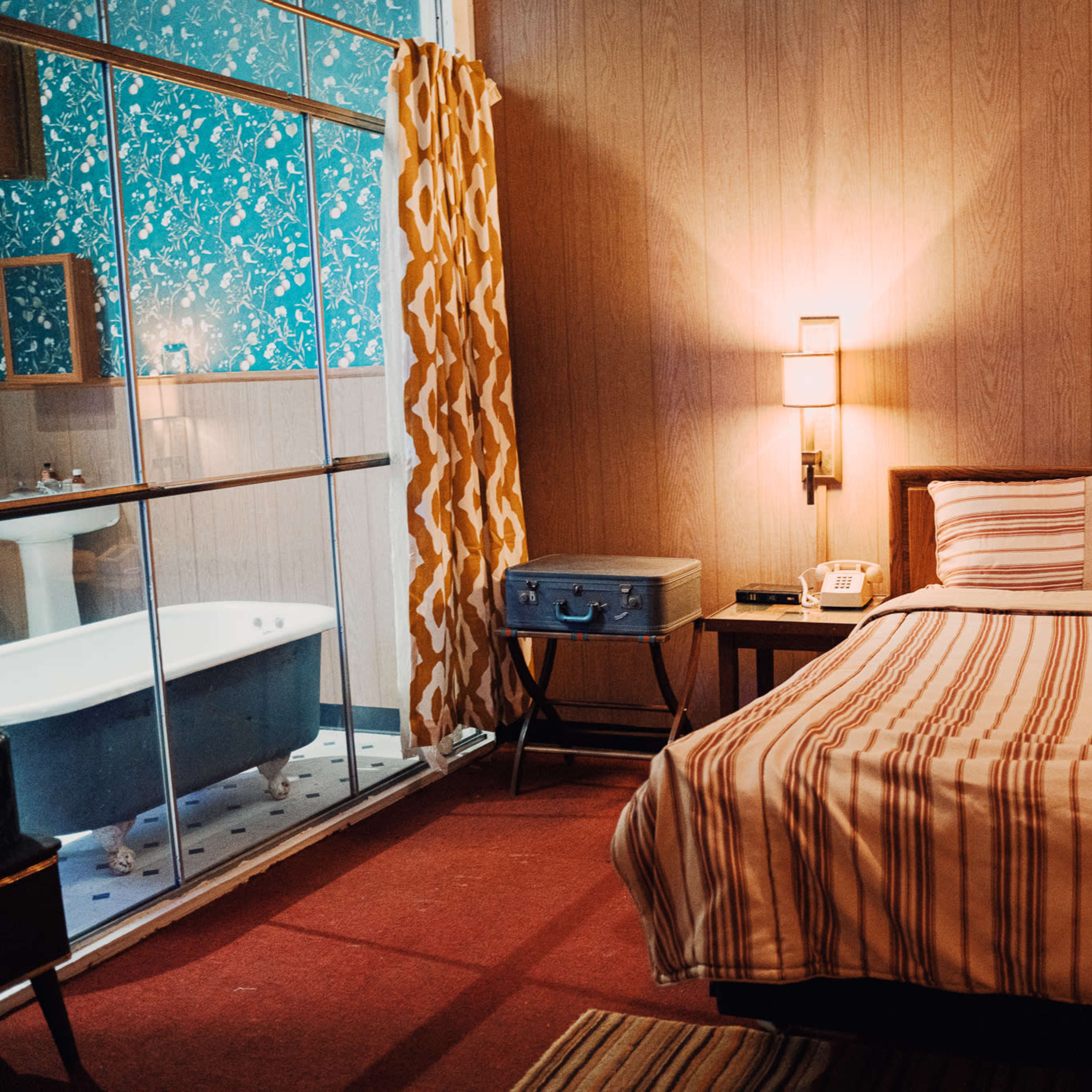 A cozy bedroom with a striped bedspread, two bedside lamps, a vintage-style telephone, and a visible bathroom area featuring a freestanding bathtub.