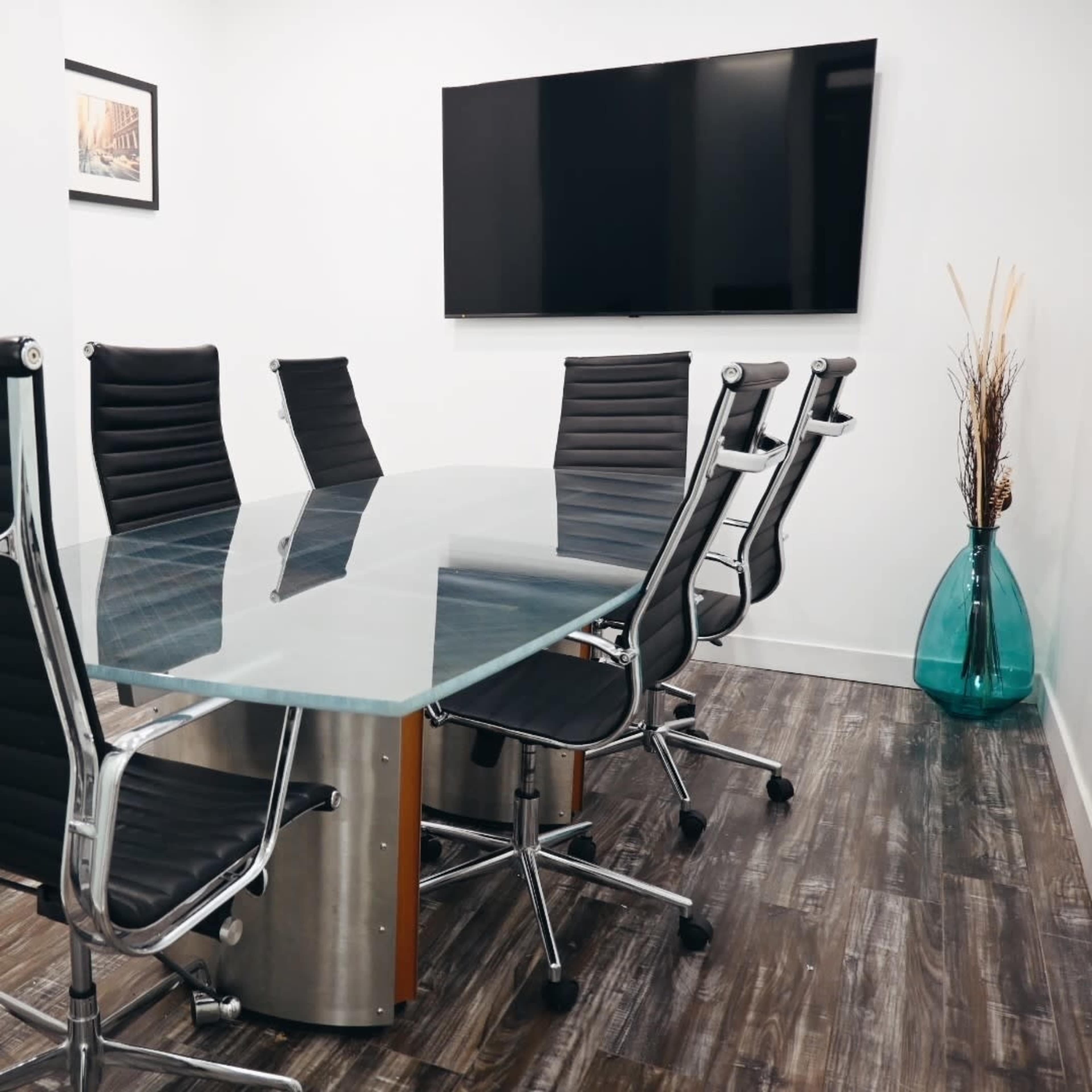 A modern conference room features a glass table surrounded by black leather swivel chairs, with a flat-screen TV mounted on the wall and a decorative vase in the corner.