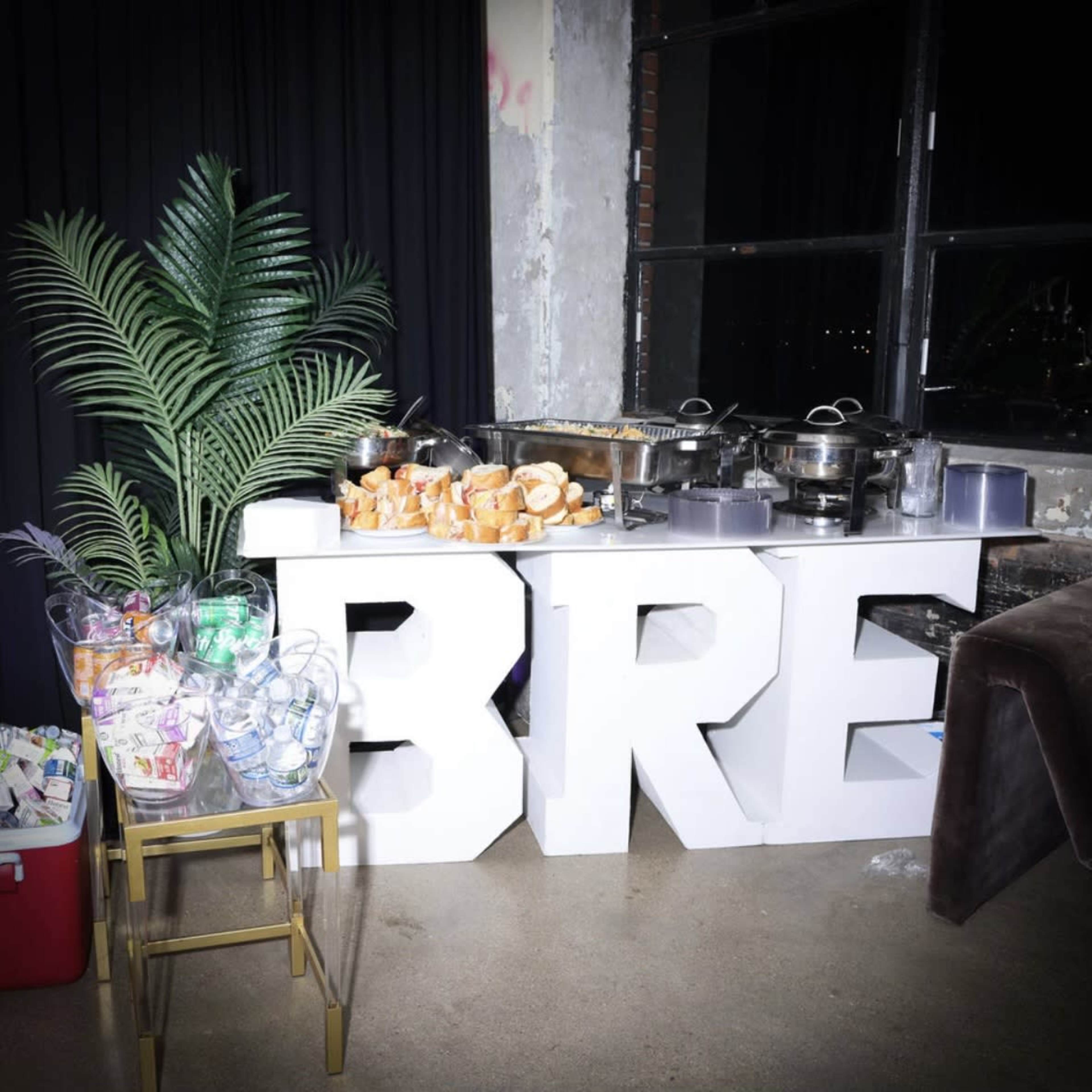 A food display featuring large letters spelling "BRE" is set up beside a decorative plant and various catering trays.