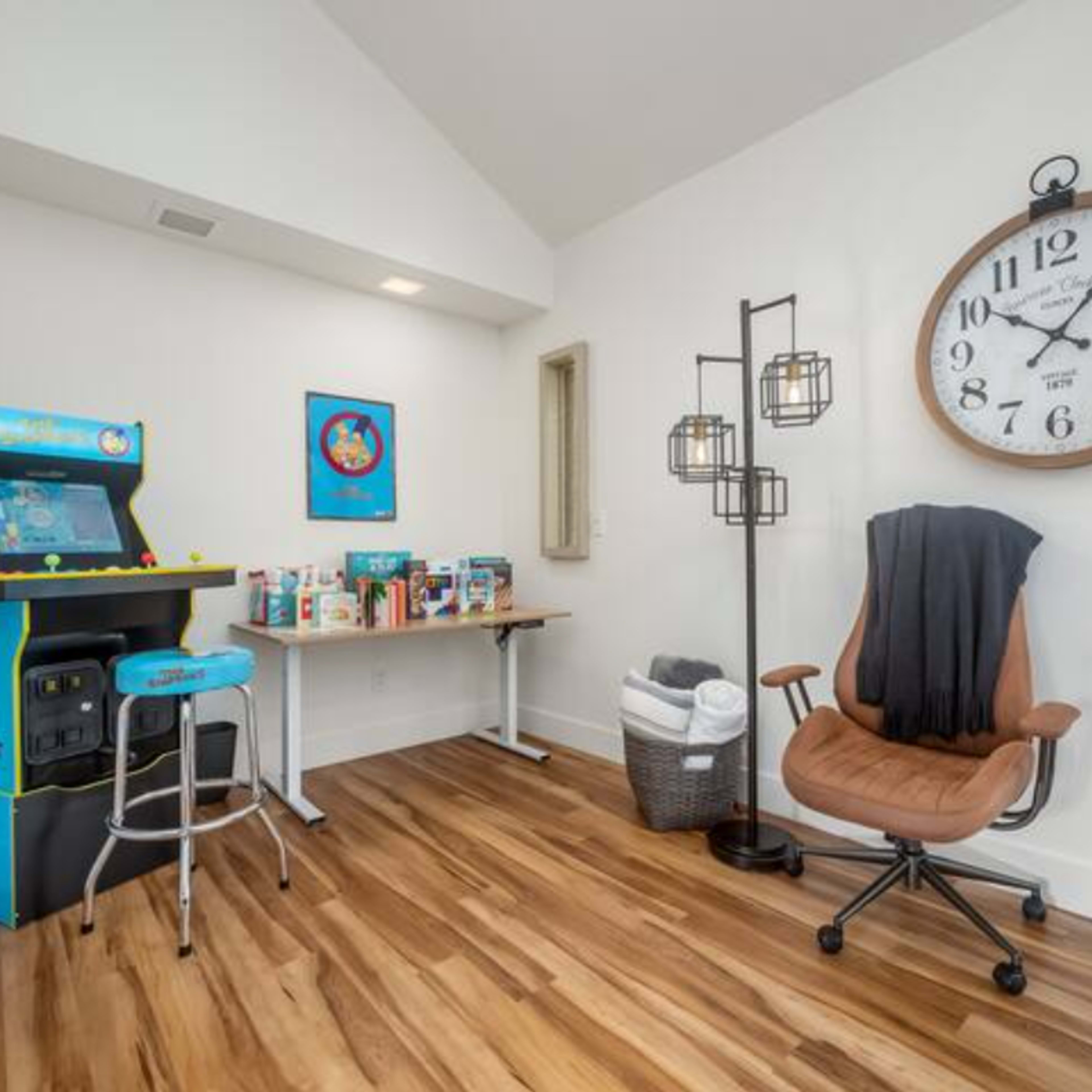 The room contains an arcade game, a clock, a chair, and a table with various items on it, all set against a backdrop of wooden flooring.