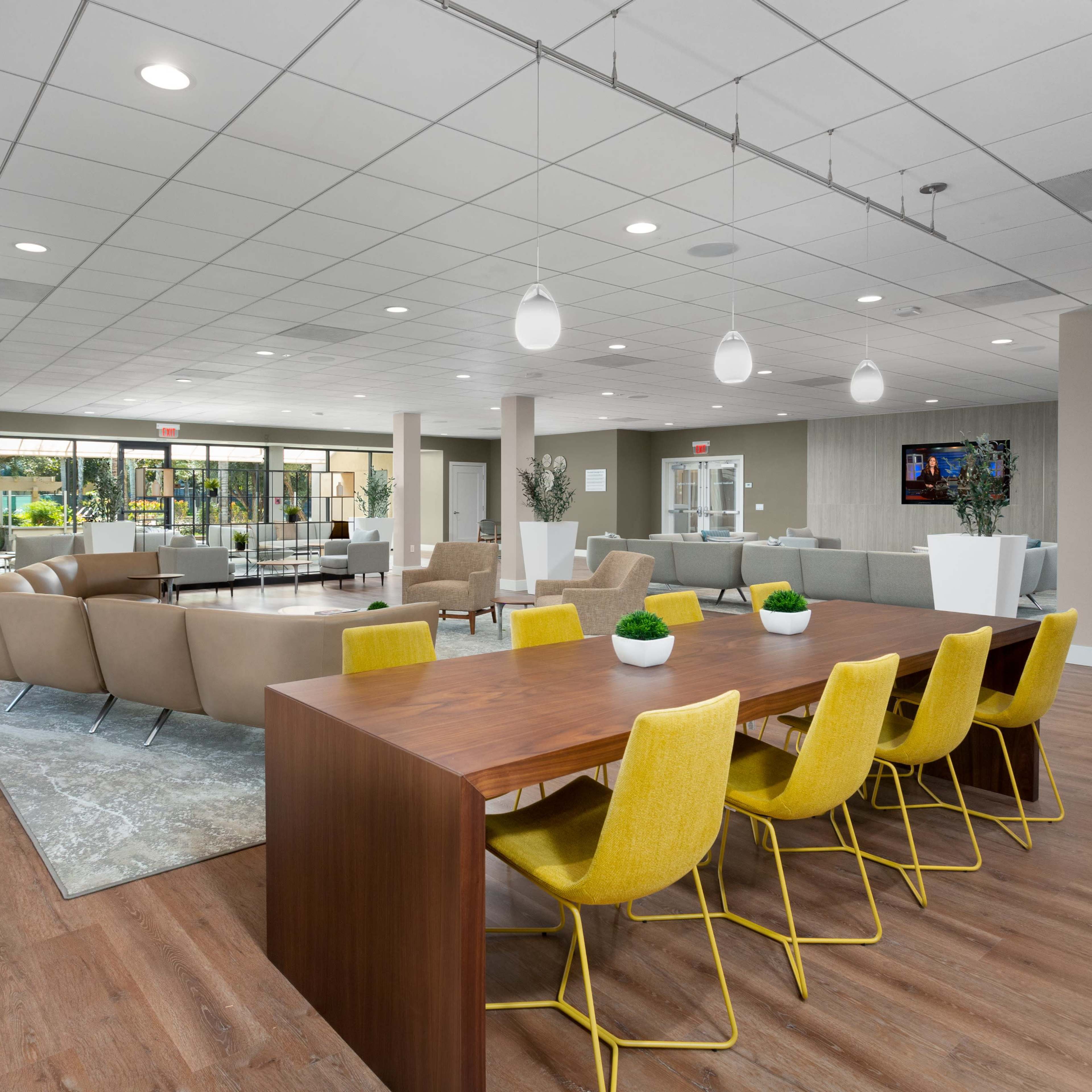 A modern interior space featuring a dining area with a long wooden table and yellow chairs, along with a lounge area that includes beige sofas, all under bright lighting.