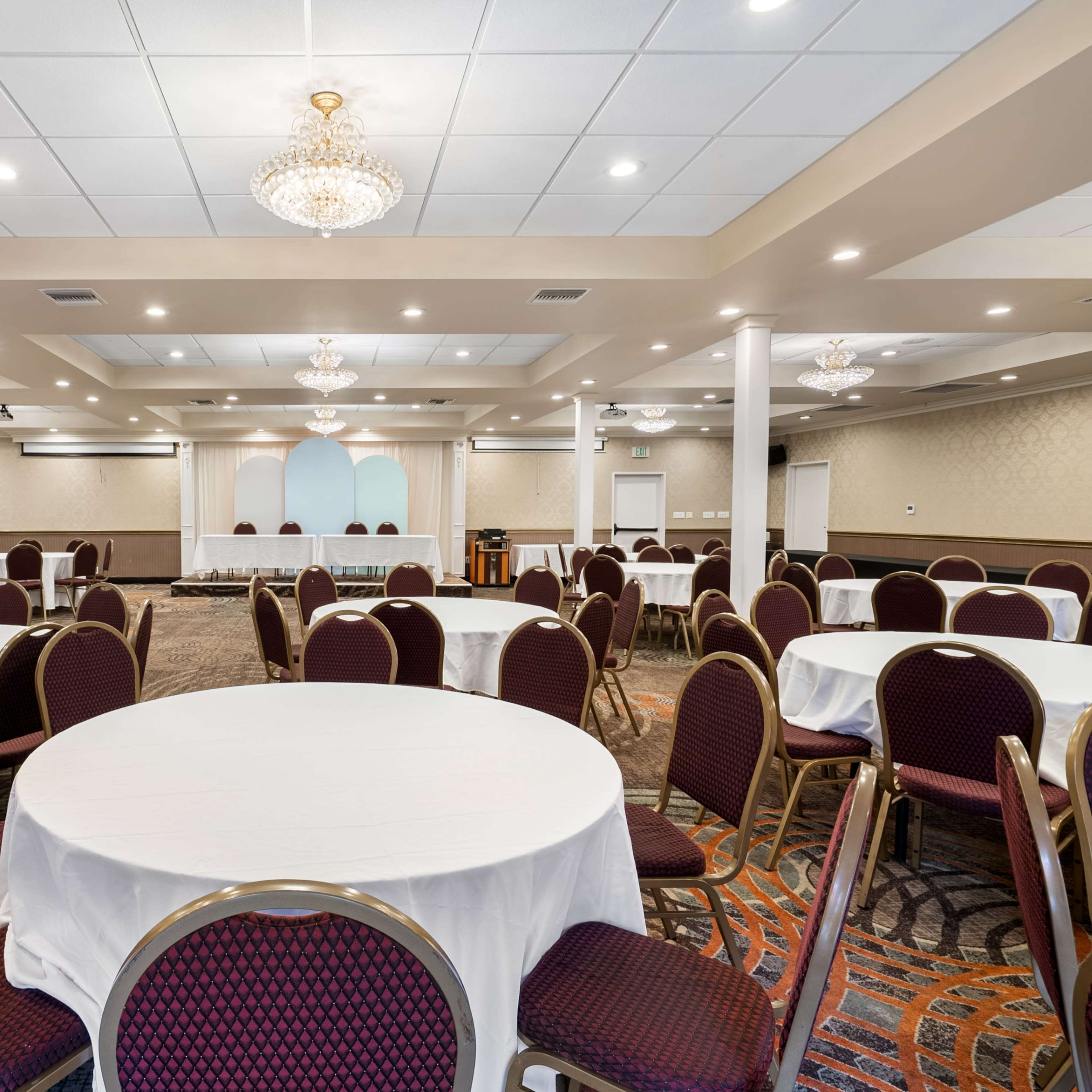 A spacious banquet hall features round tables with white tablecloths and elegant chandeliers overhead.