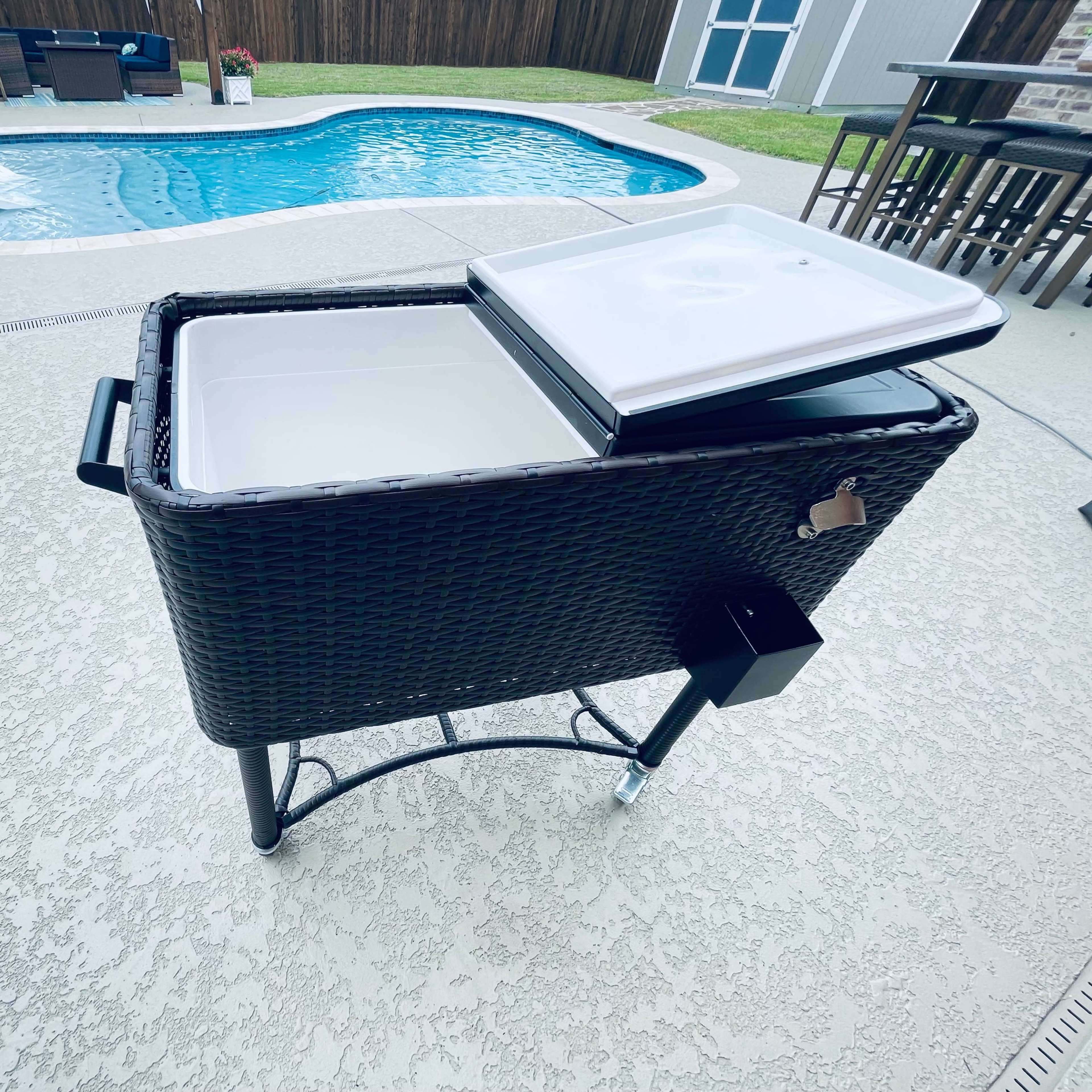 A rectangular wicker cooler with a lid sits beside a swimming pool on a textured patio.
