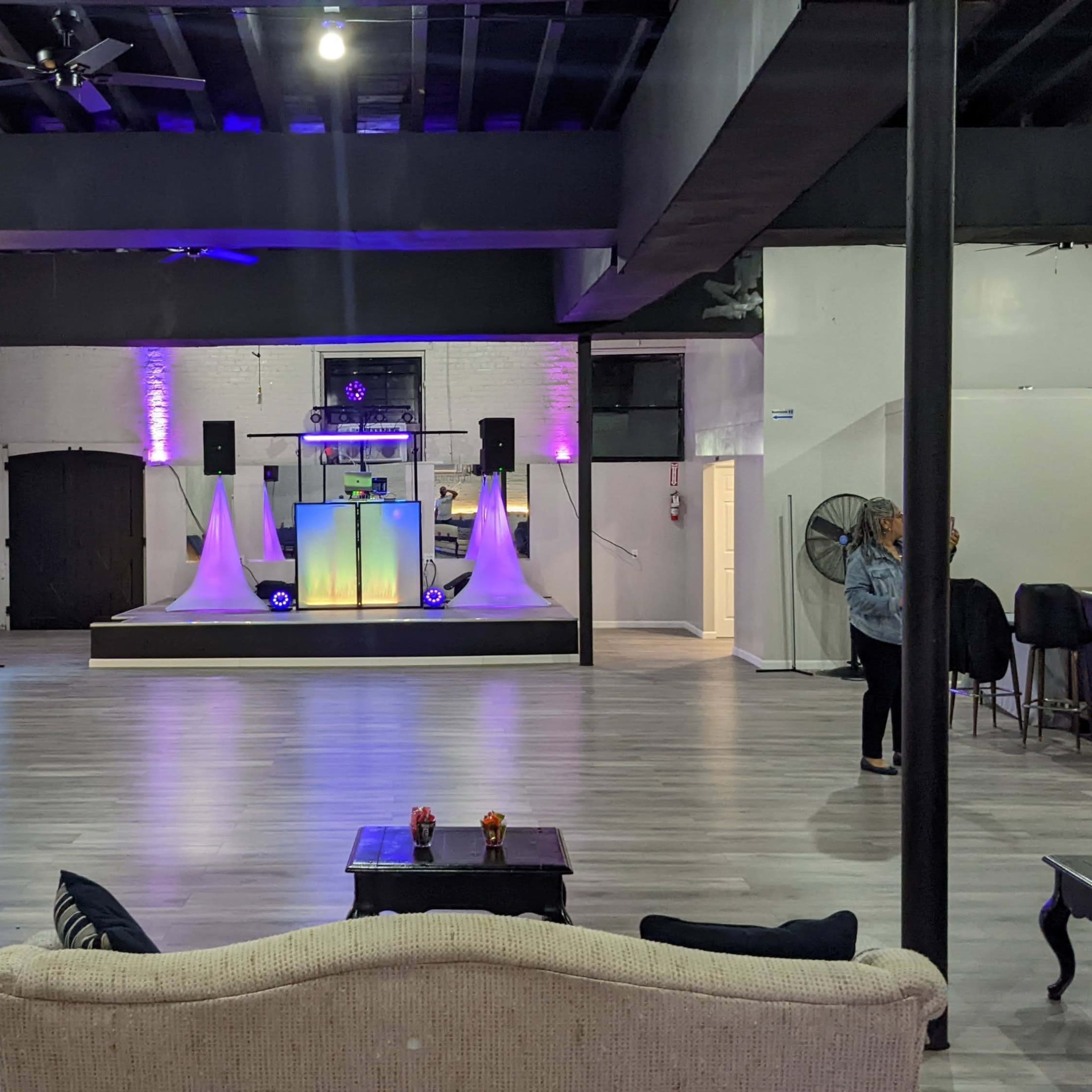 A spacious event venue with a stage featuring speakers and decorative lighting, surrounded by seating areas and a bar in the background.