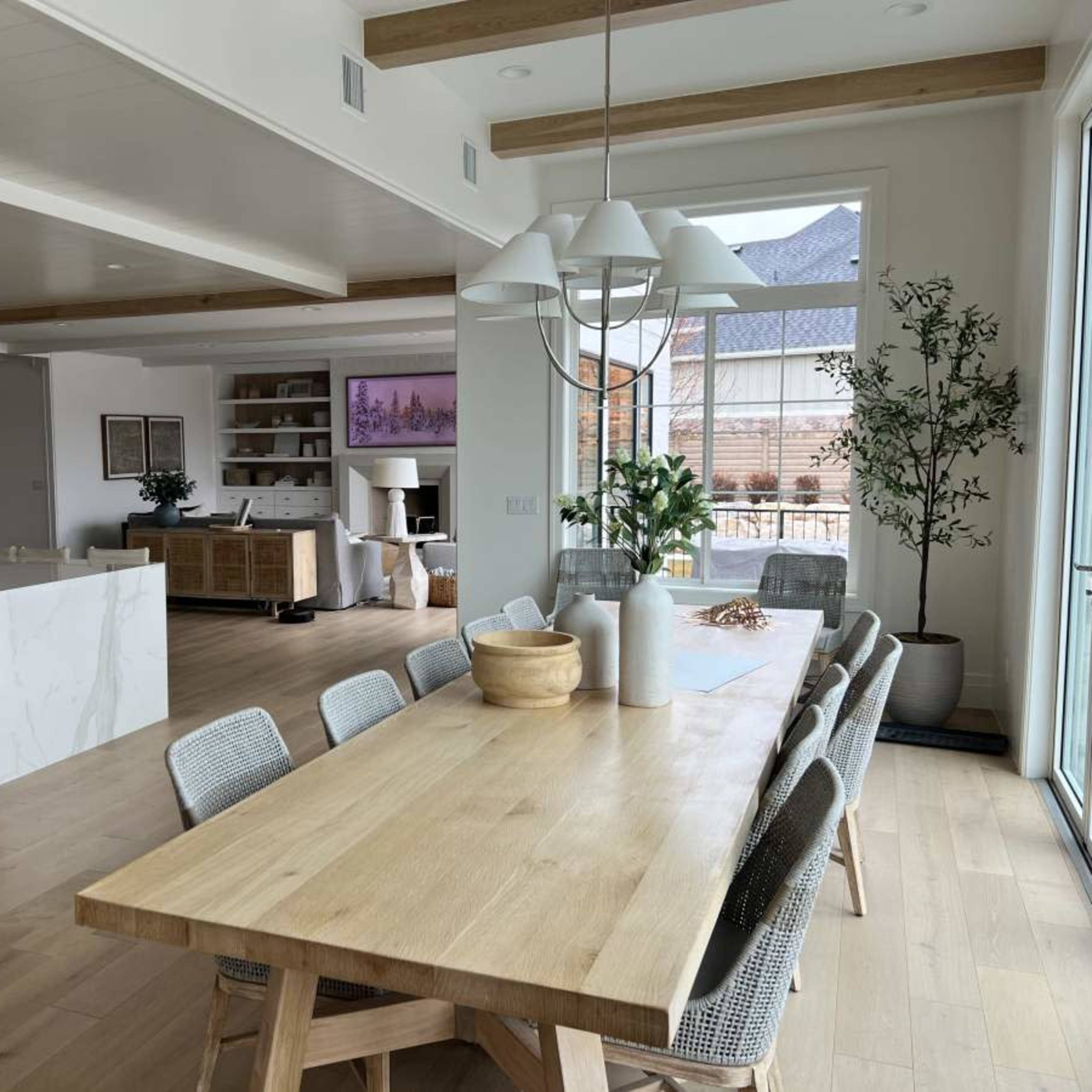 A modern dining area features a long wooden table surrounded by chairs, with an open kitchen and living space in the background.