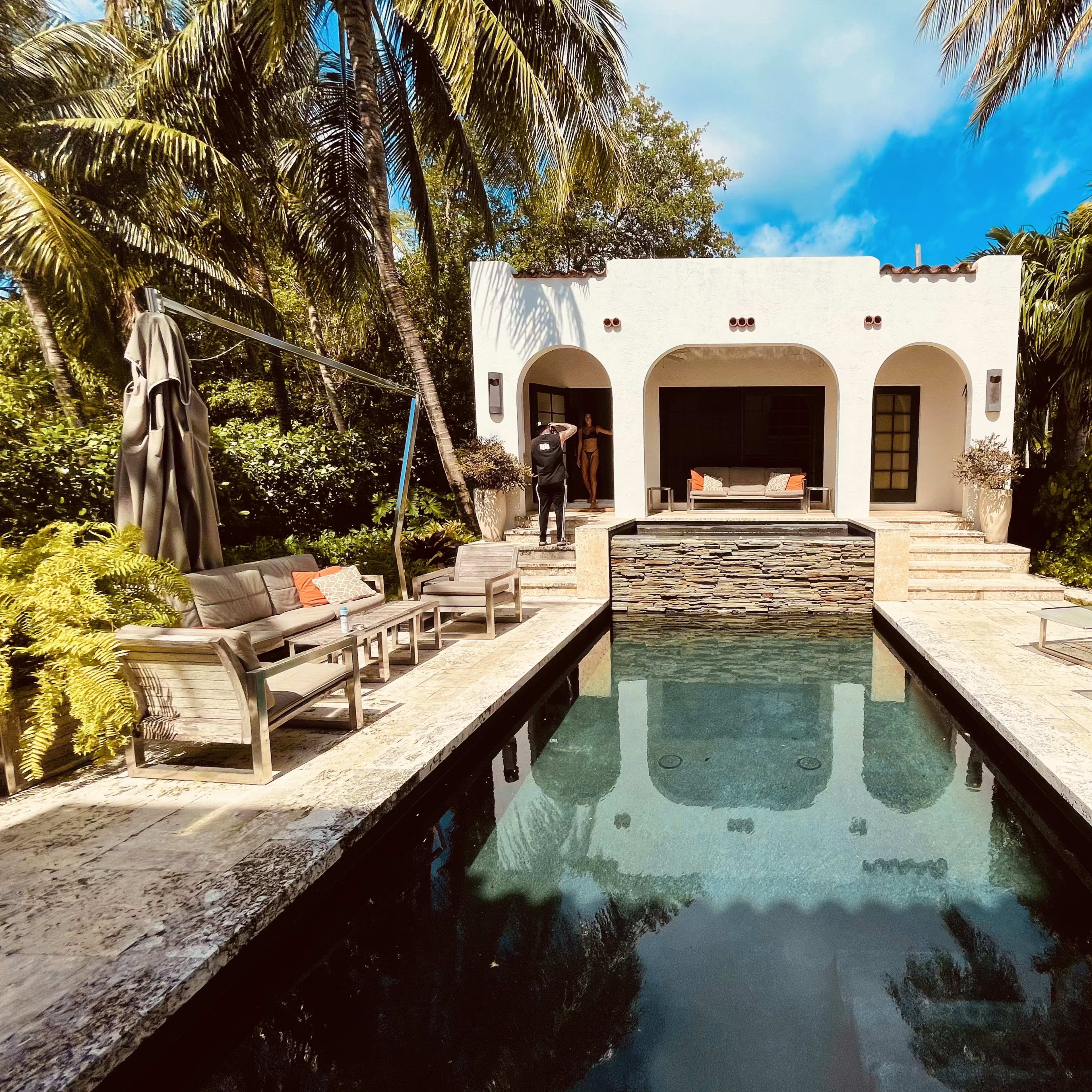 A serene outdoor scene features a rectangular swimming pool surrounded by palm trees, lounge chairs, and a small modern pavilion with a stone accent wall.