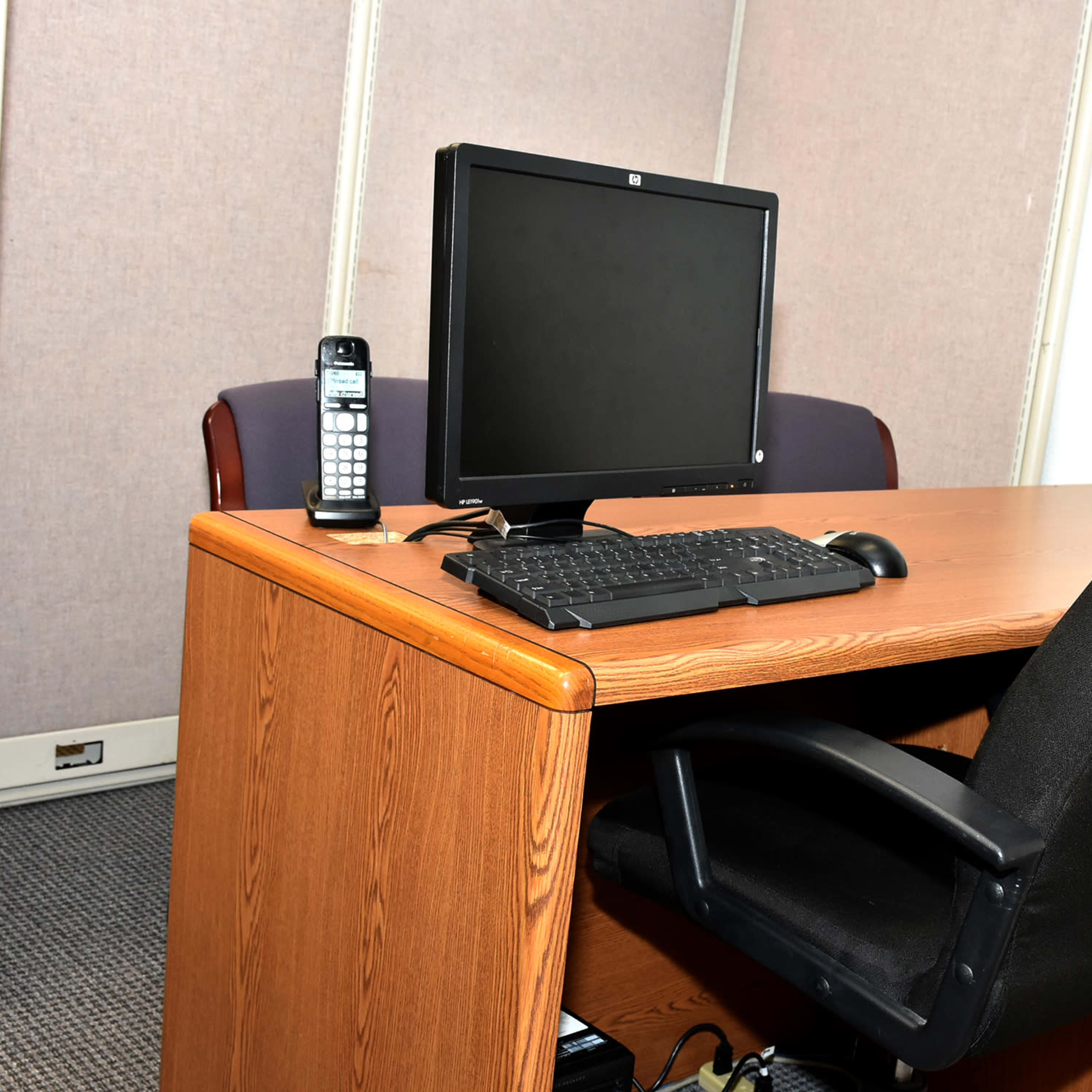 An office setup with a wooden desk, an empty black office chair, a desktop computer monitor, a keyboard, and a telephone.