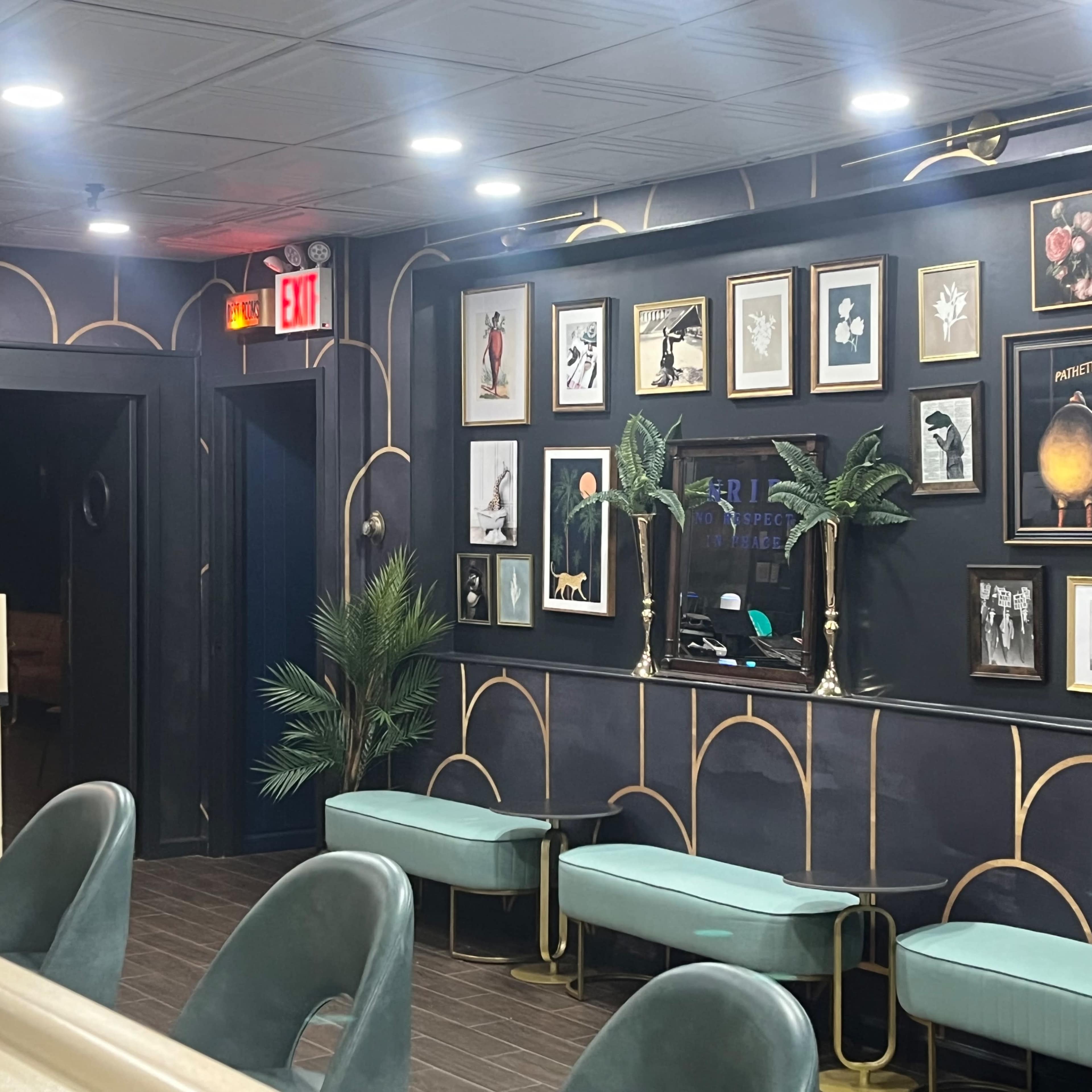 A modern waiting area featuring green upholstered seating, decorative wall frames with artwork, and a potted plant against a dark patterned wall.