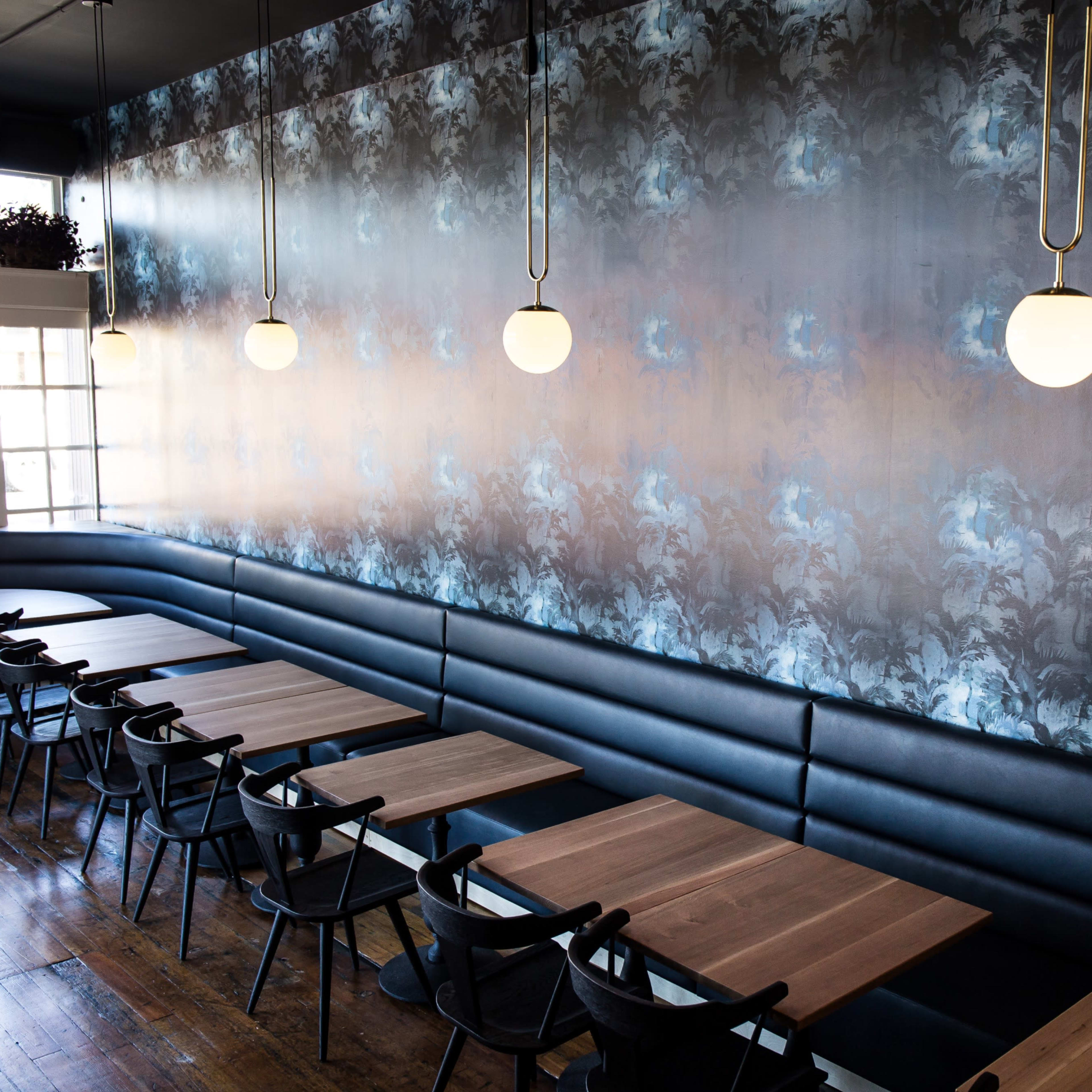 A dining area with a patterned wallpaper, wooden tables, and black seating along the walls, illuminated by round pendant lights.