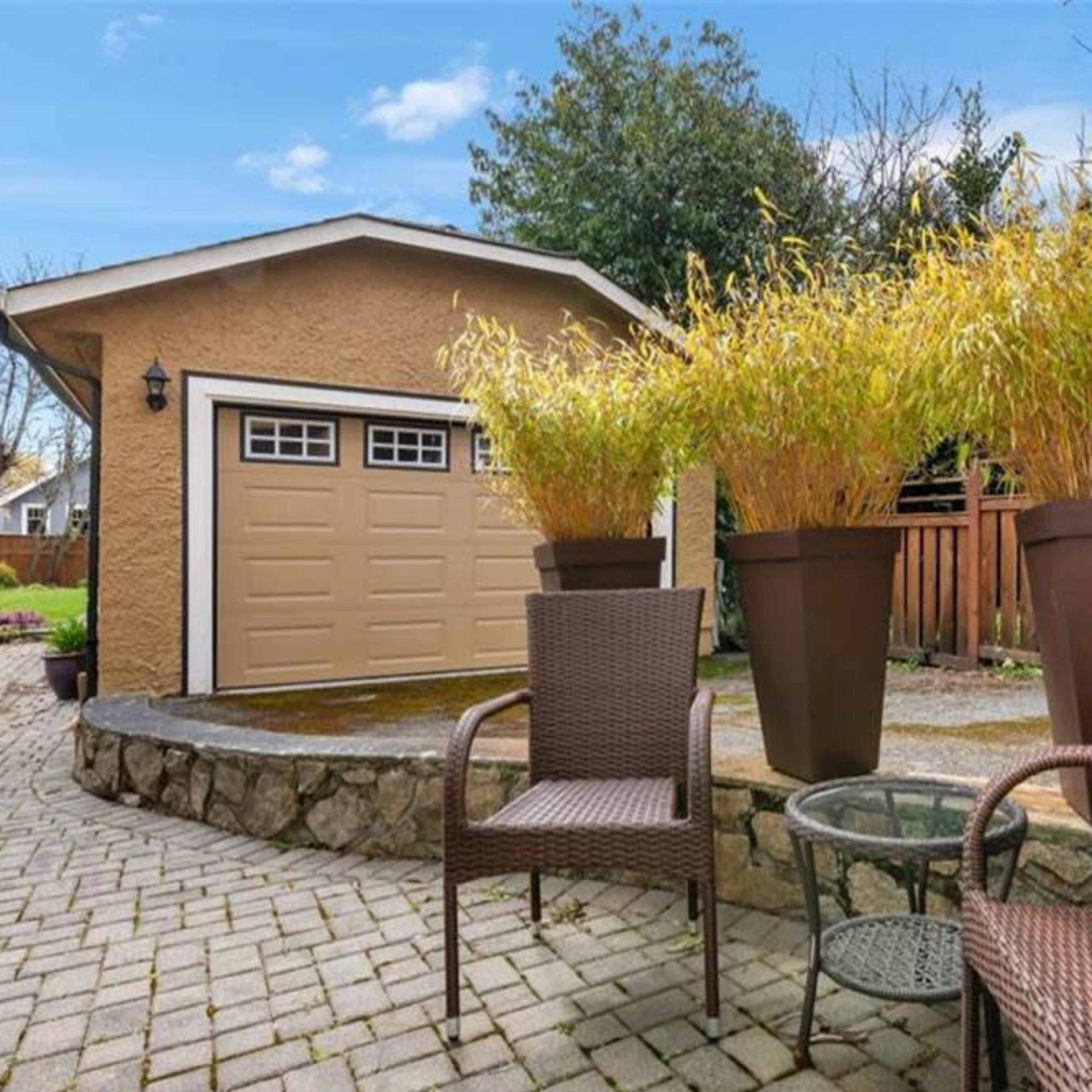 A cozy outdoor space featuring two chairs in the foreground, a stone patio, and a garage with a beige exterior in the background, surrounded by tall potted plants.