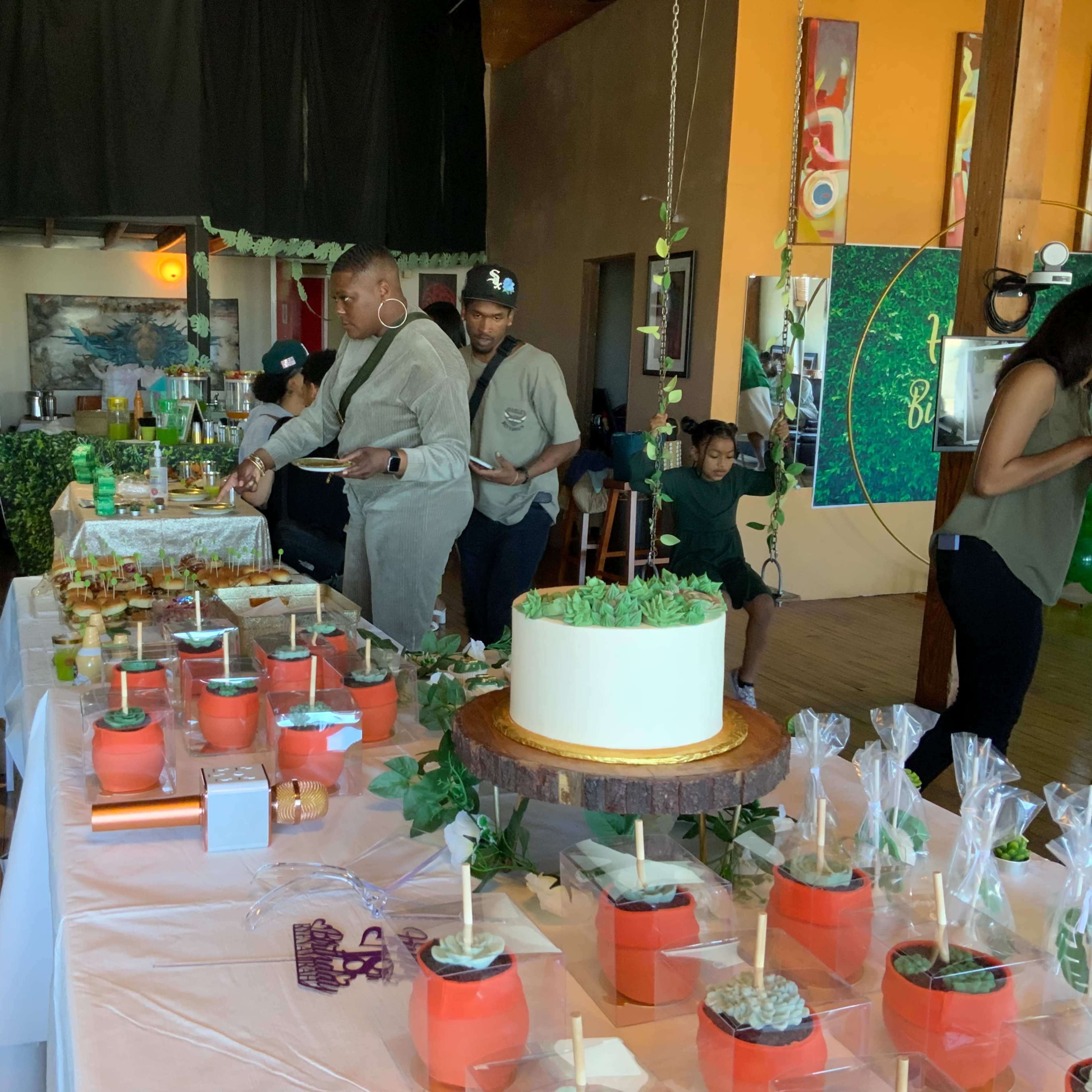 A gathering features a buffet table filled with various food items and a large cake, with guests enjoying the offerings in a decorated indoor space.