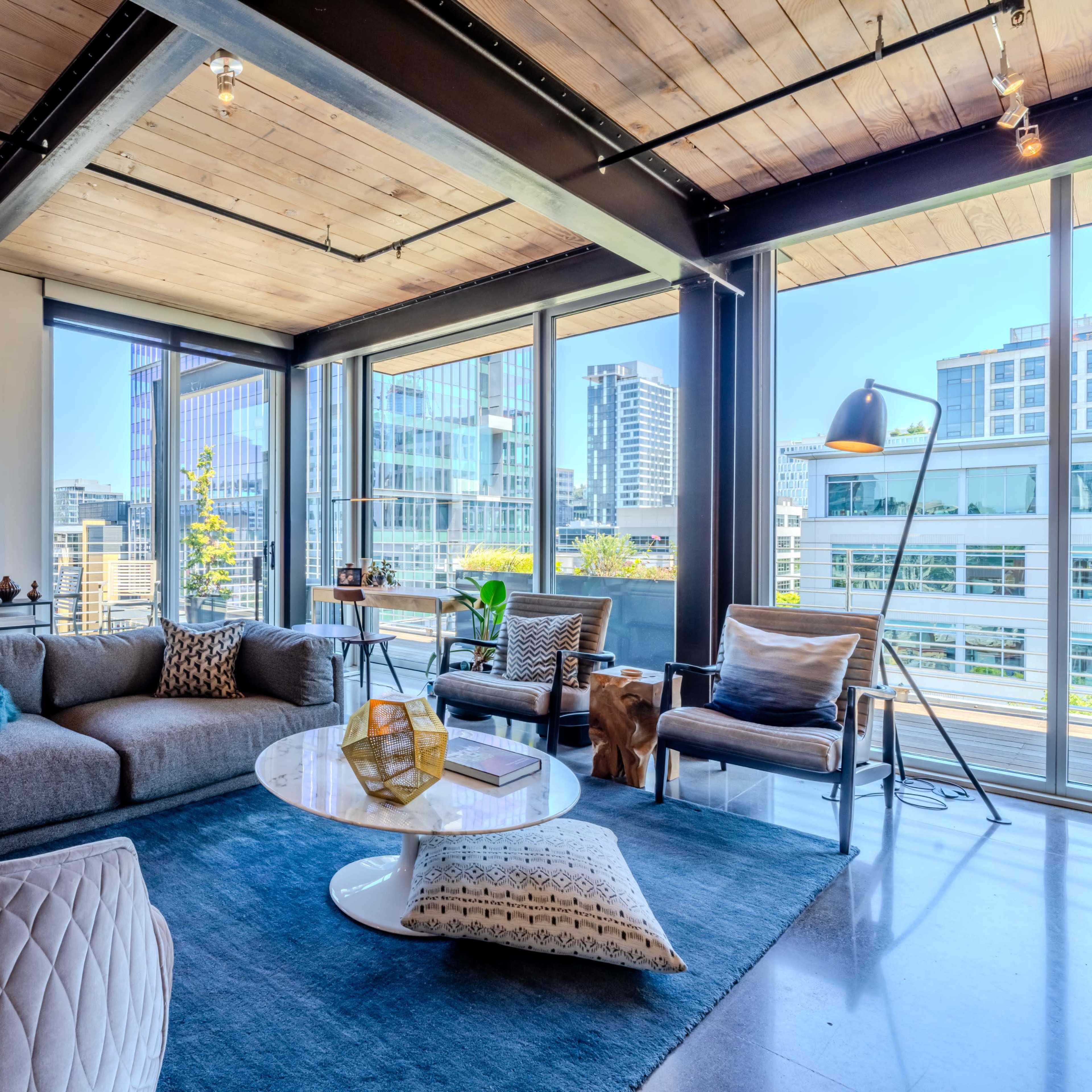 A modern living room features a gray sectional sofa, a round coffee table, and large windows showcasing a city skyline.