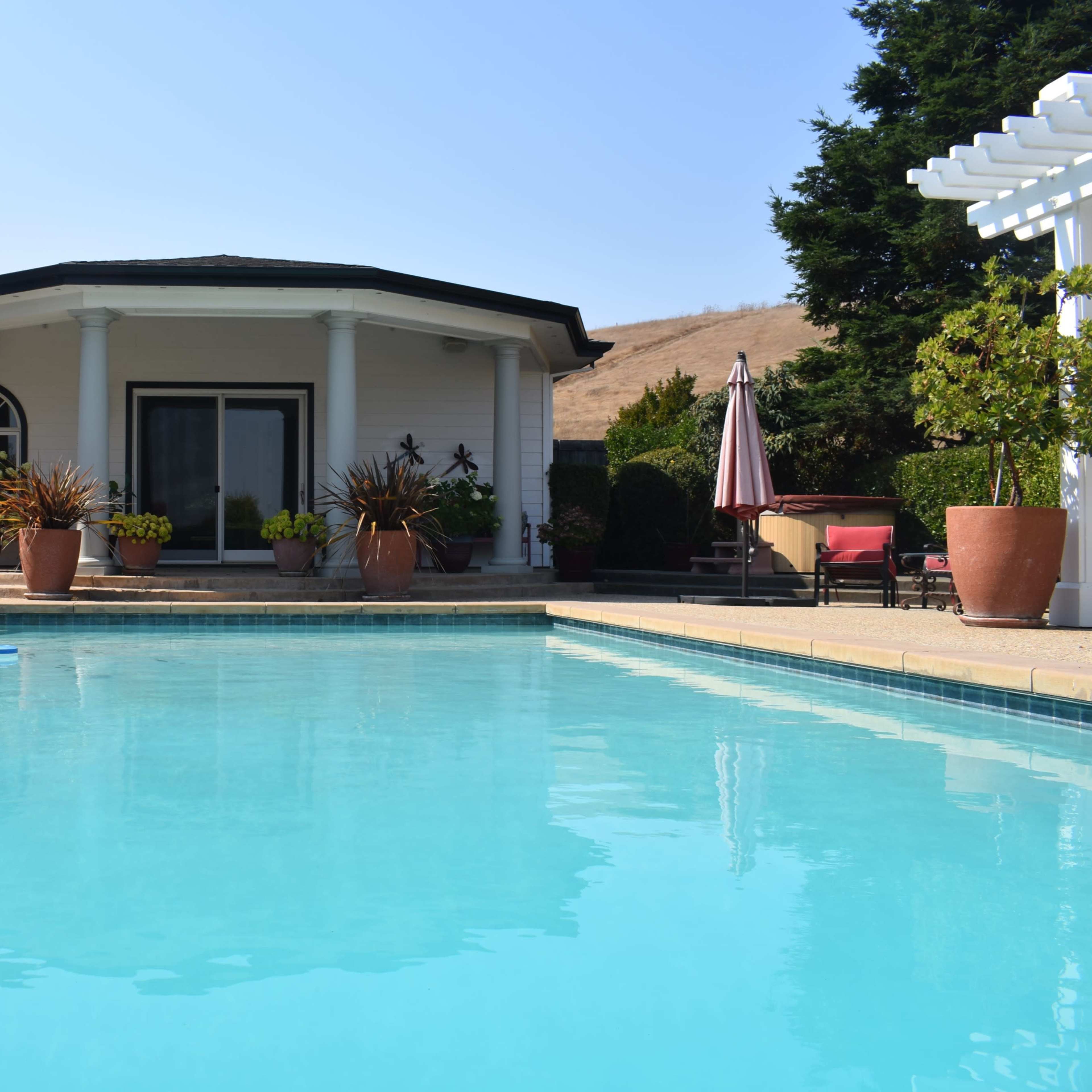 A clear blue swimming pool reflects the surrounding patio and a house with pillars and a circular roof.