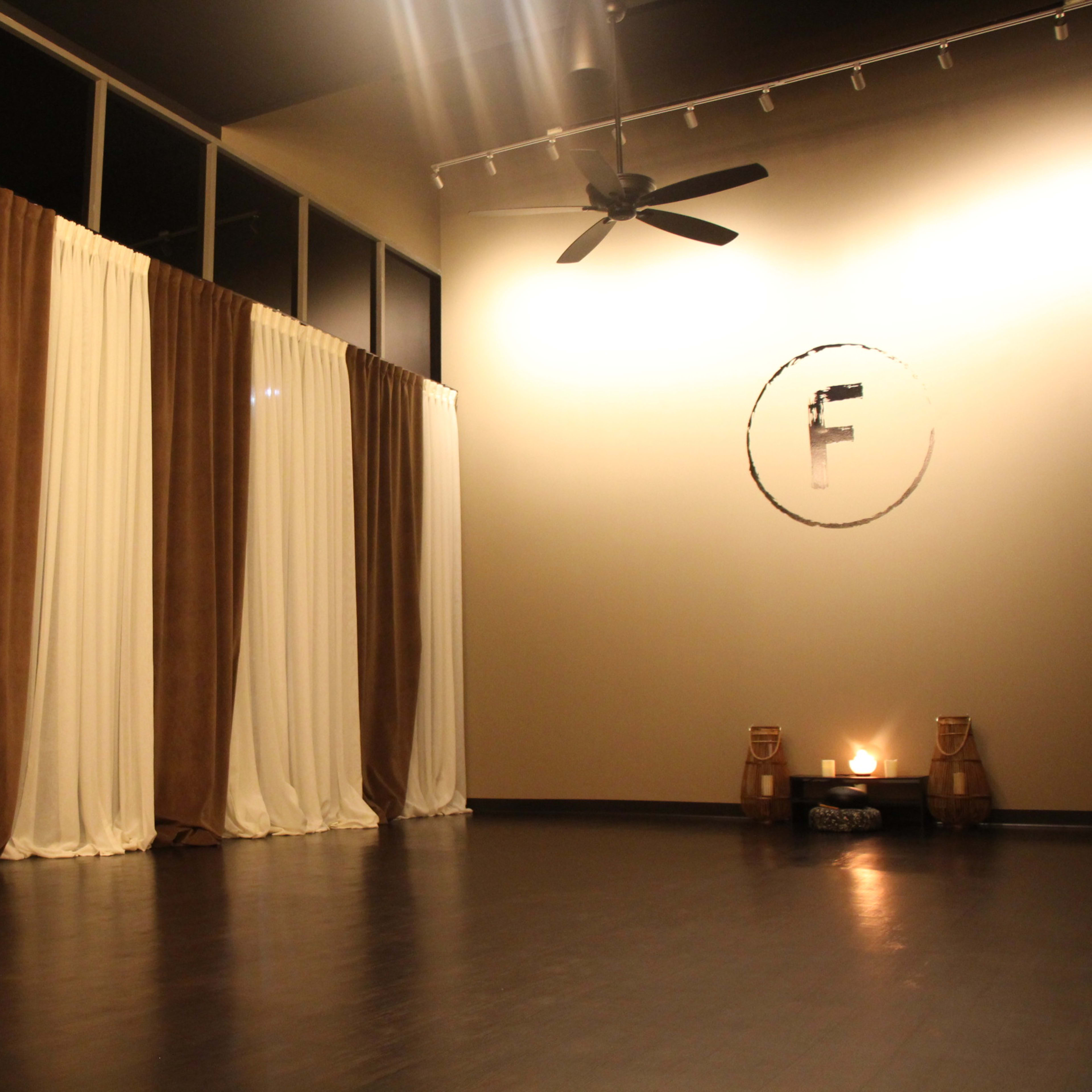 A spacious room with large, floor-to-ceiling curtains in brown and white, a fan on the ceiling, and a simple circular wall decoration with an "F" in the center, along with two candles on a table.